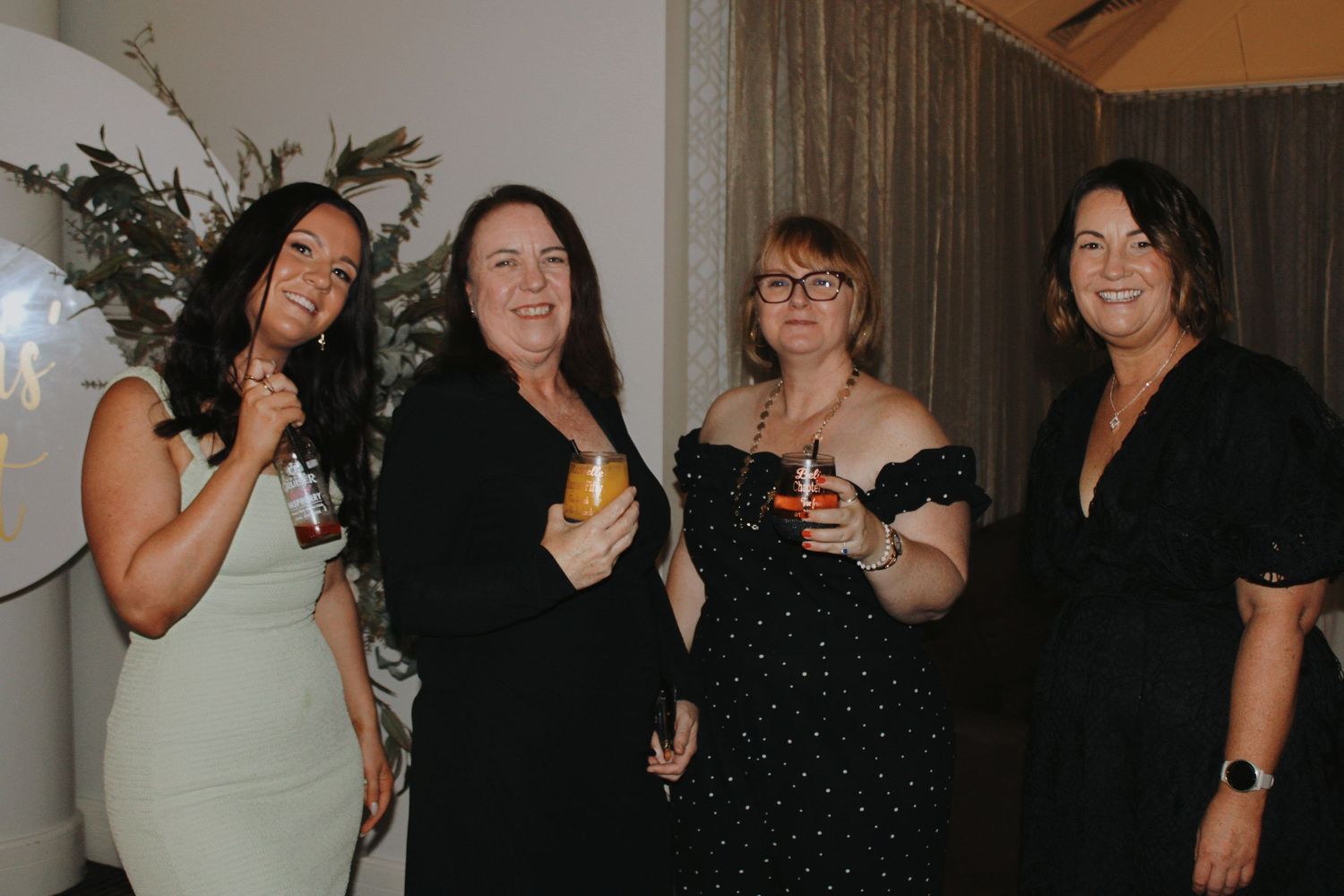 Four women smiling and holding drinks together at an indoor social celebration.