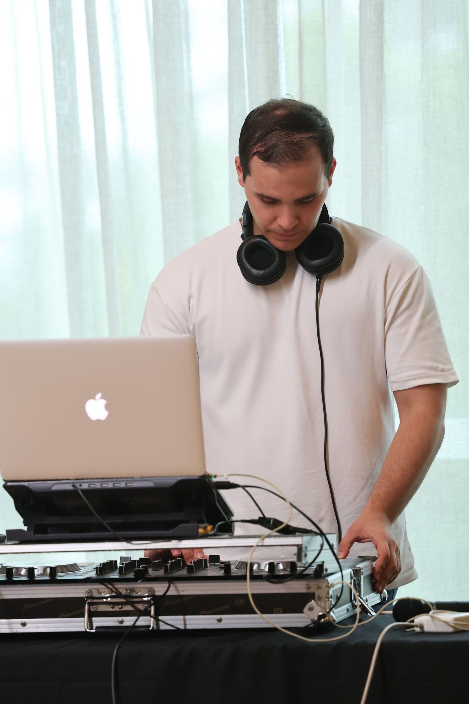 Male DJ wearing headphones around neck working on Apple laptop and mixing equipment at an event.