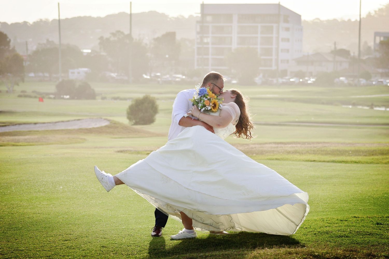 Groom dips bride for a romantic kiss on a green golf course, her white dress flowing in the breeze.