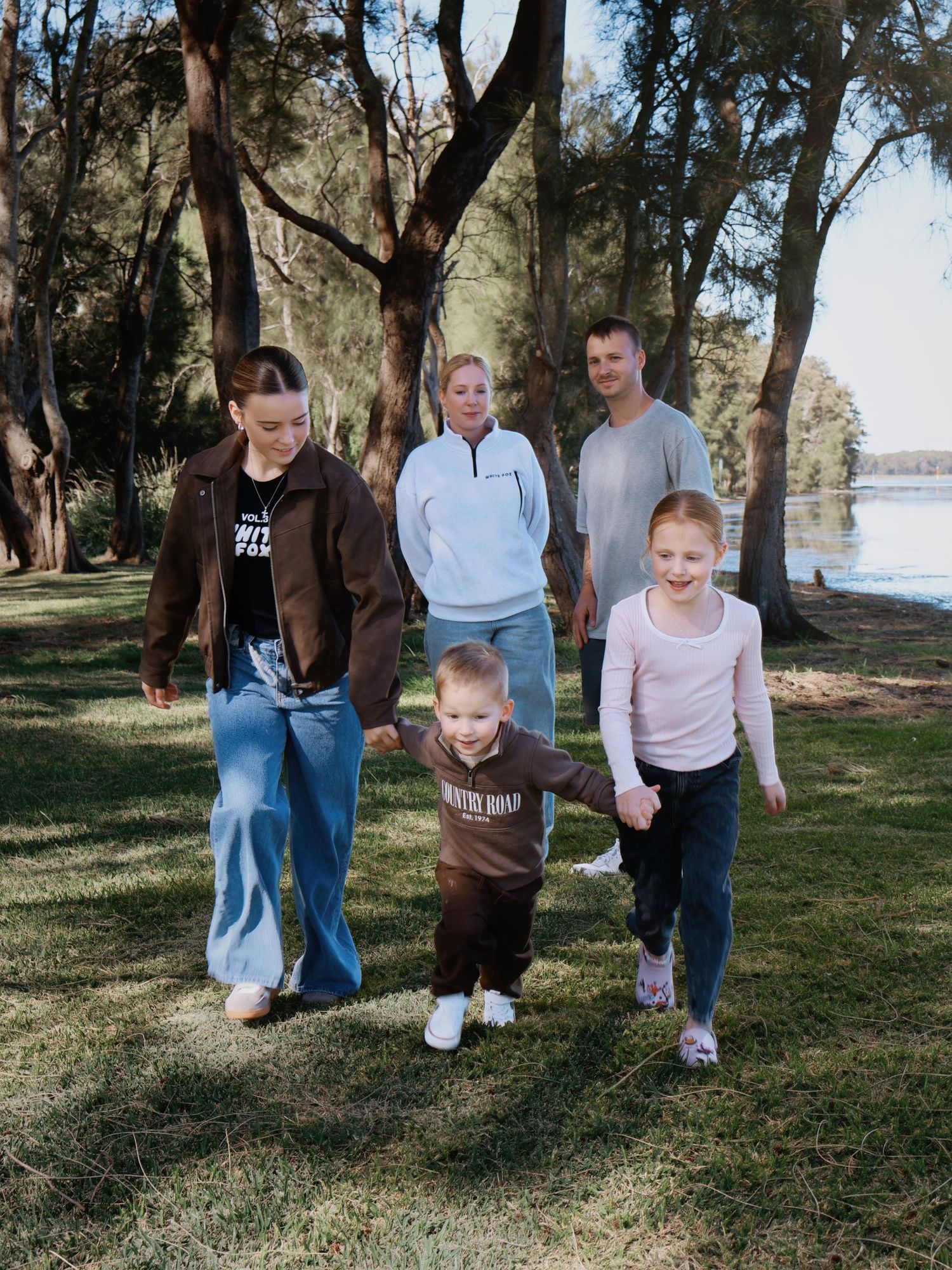 A happy family of five walks together through a tree-lined park near a lake on a sunny day.