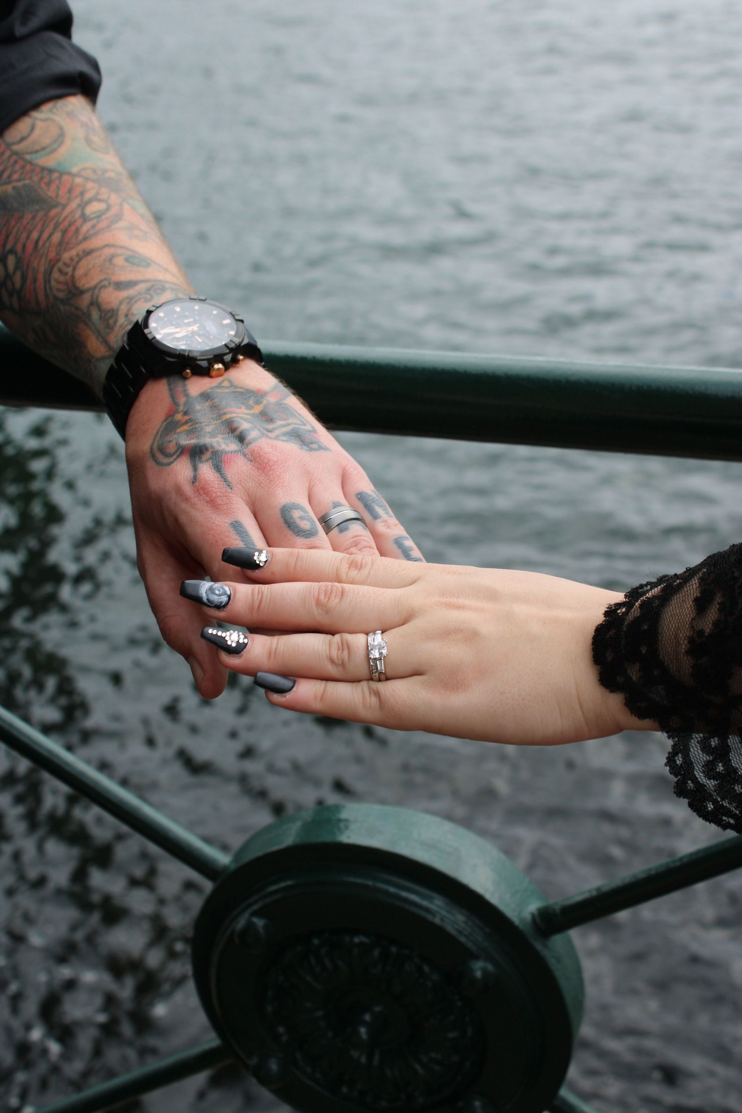 Tattooed groom hand holding bride's hand with decorative nails and rings by waterside railing.