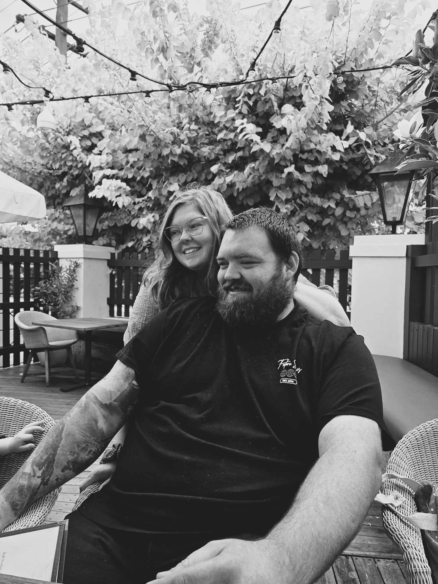 A bearded man and smiling woman pose together on an outdoor patio with string lights and lush greenery.