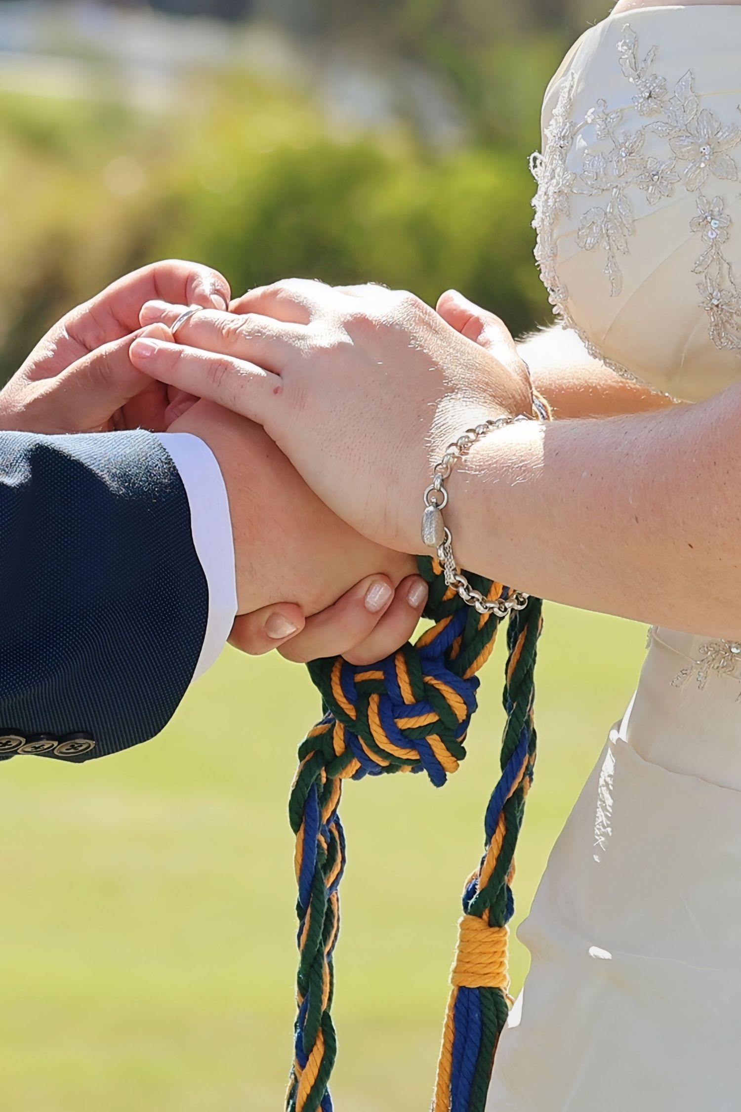 Bride and groom hands bound together with colorful green, gold, and blue rope during a handfasting wedding ceremony.