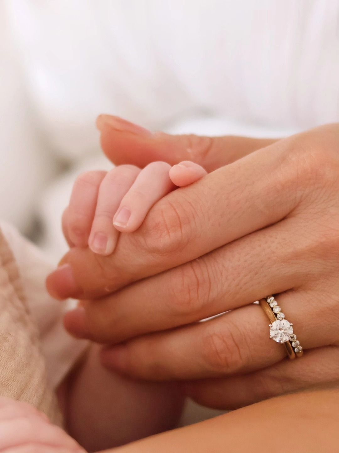 Close-up of a newborn baby holding an adult finger, with a diamond engagement and wedding ring visible.