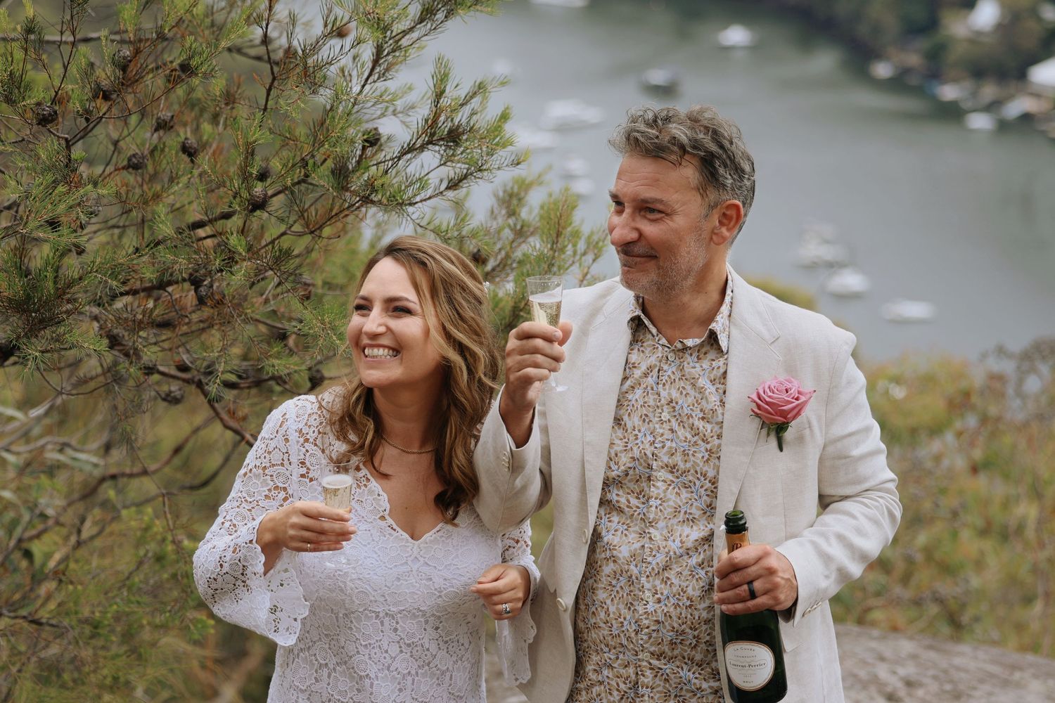 Happy couple celebrating outdoors with champagne glasses and wine bottle overlooking a scenic bay.