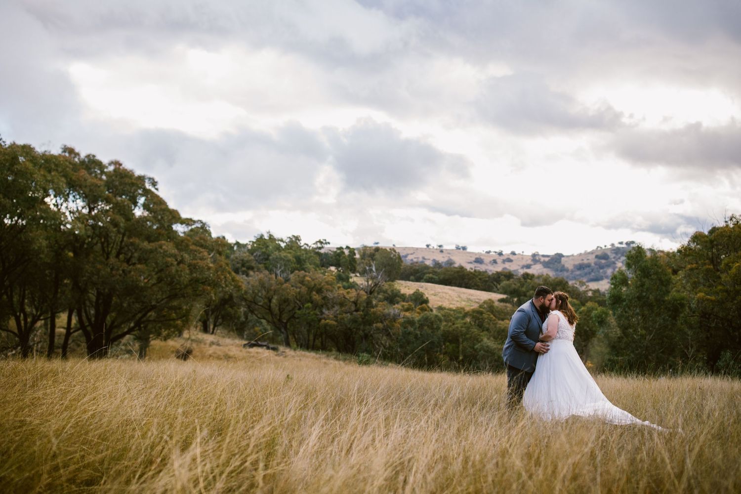 Bride and groom share a romantic moment in a golden field with rolling hills and dramatic cloudy sky backdrop.