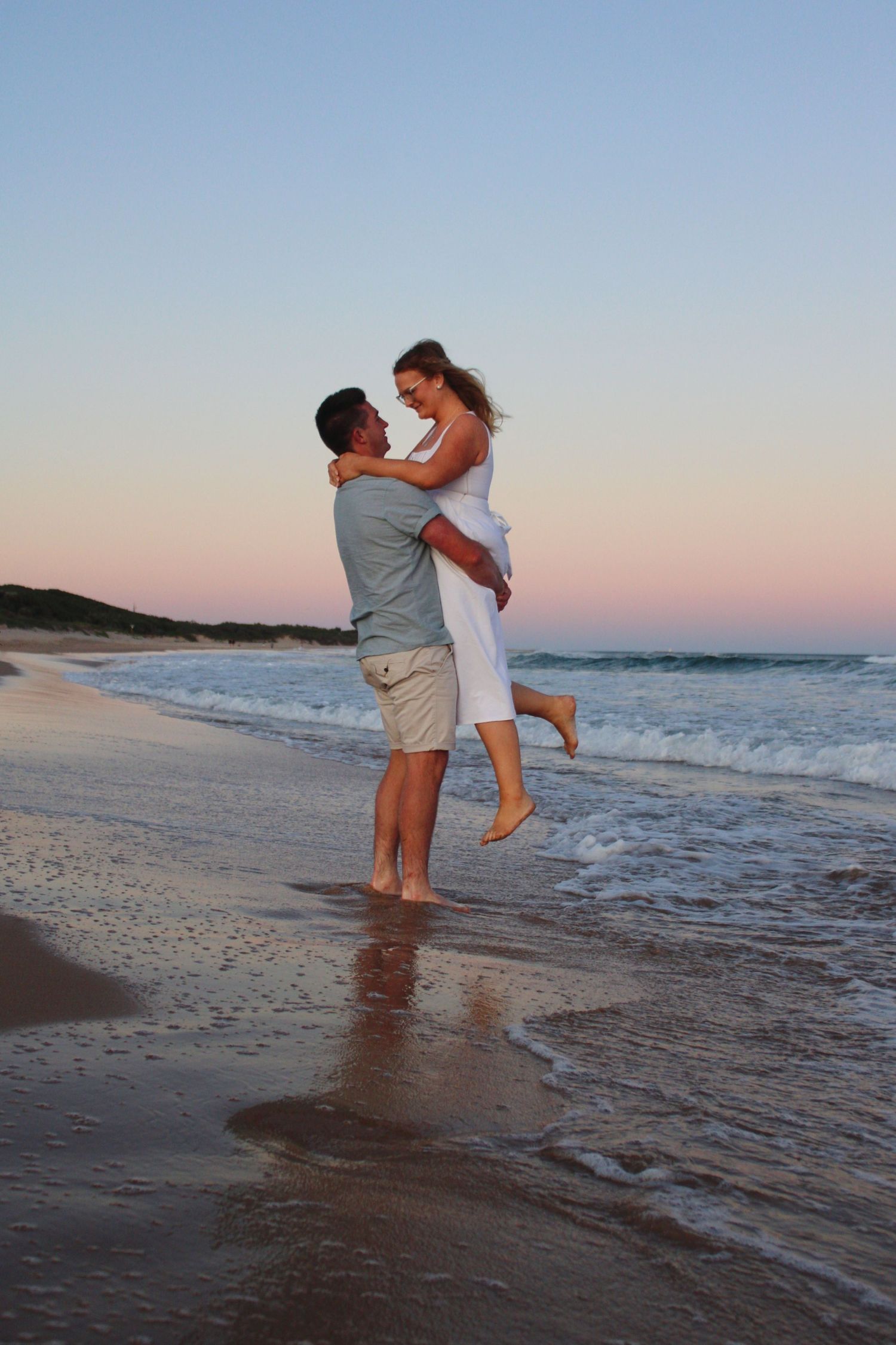 Man lifting woman in white dress on a sandy beach at sunset with ocean waves behind them.