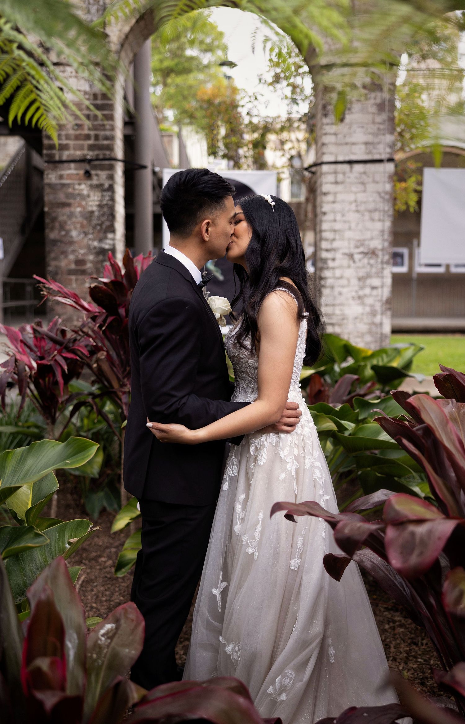Bride and groom sharing a kiss surrounded by lush tropical plants under stone archway.