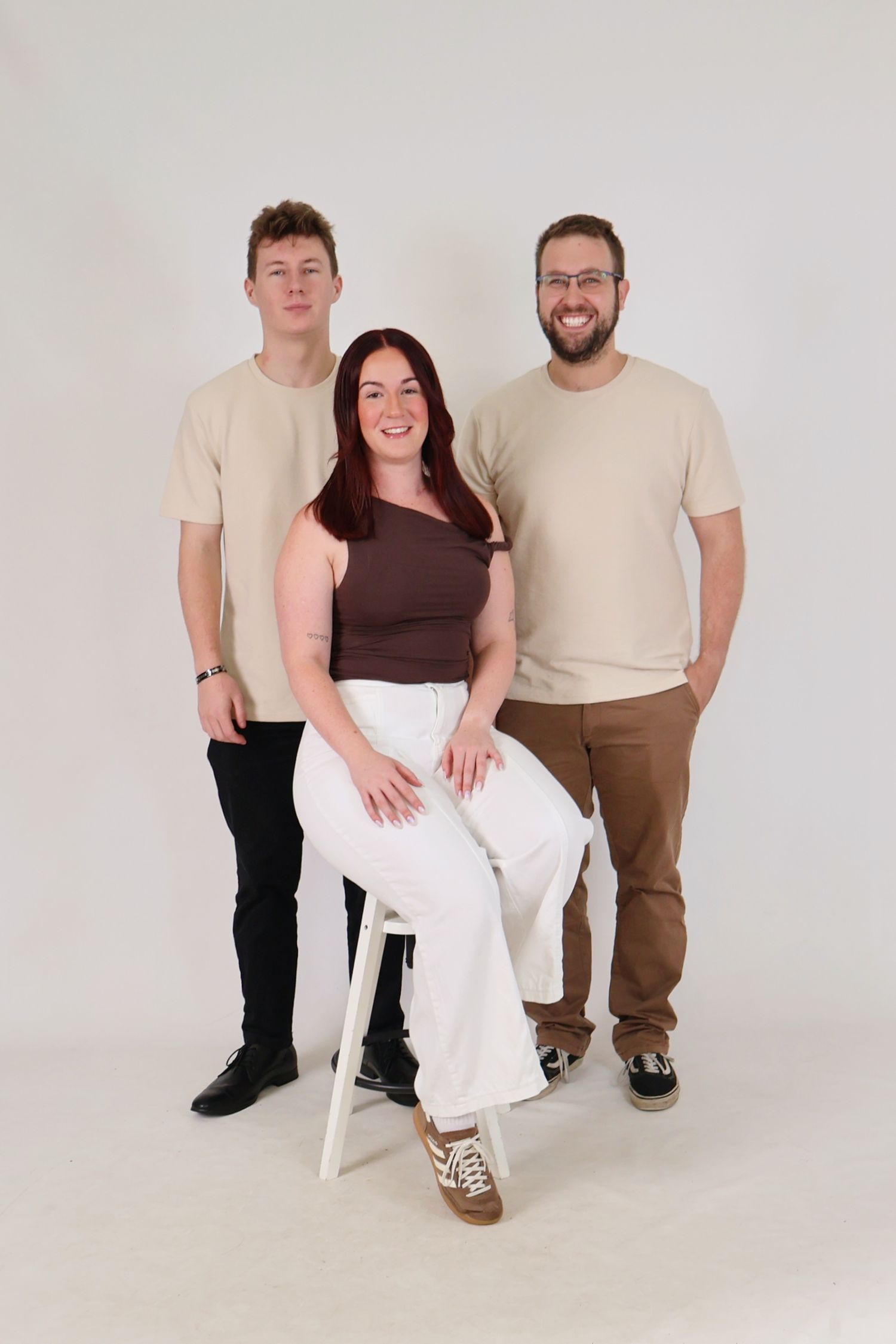 Woman seated in white pants and brown top poses with two men in beige shirts in a studio.