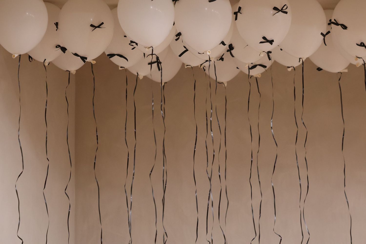 White helium balloons with black ribbon strings floating against a beige wall at an indoor party.
