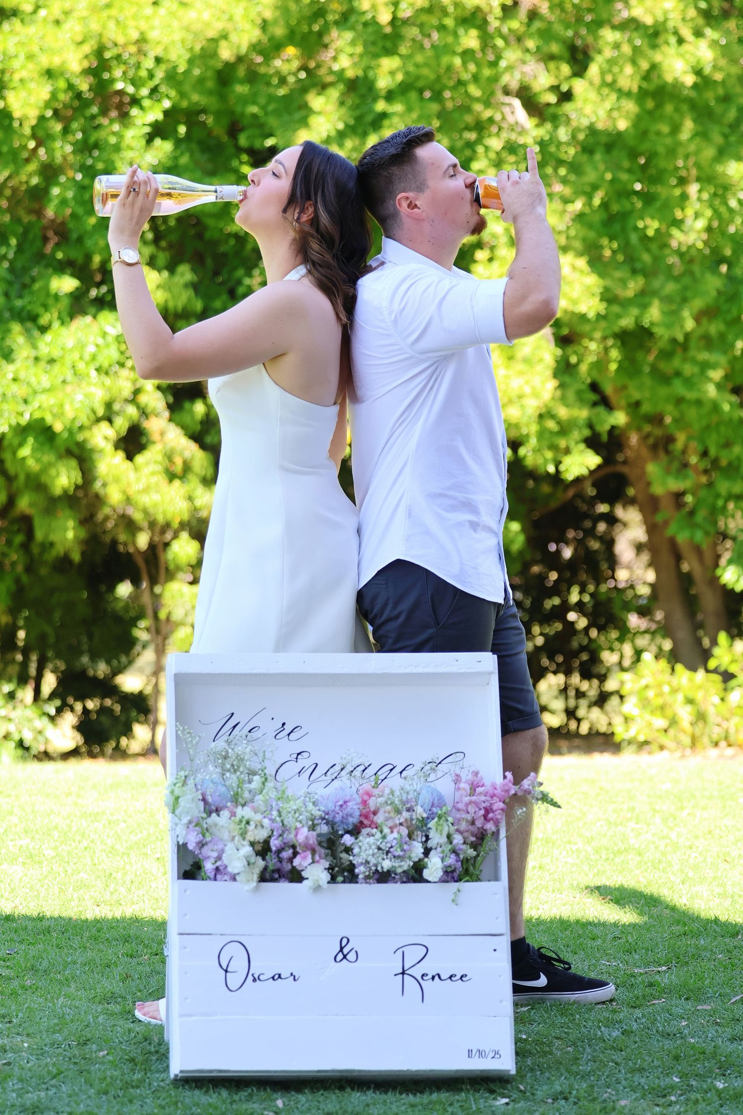 Engaged couple drinking champagne back-to-back beside a floral We're Engaged box reading Oscar and Renee.