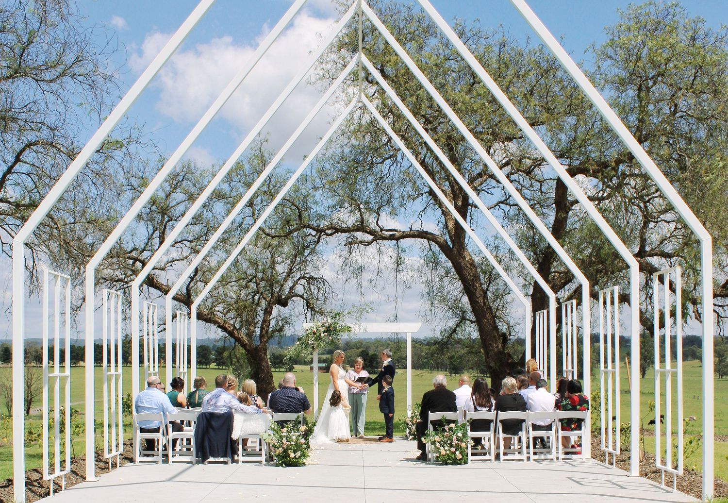 Elegant outdoor wedding ceremony under white geometric arch frames with guests seated on a sunny day.