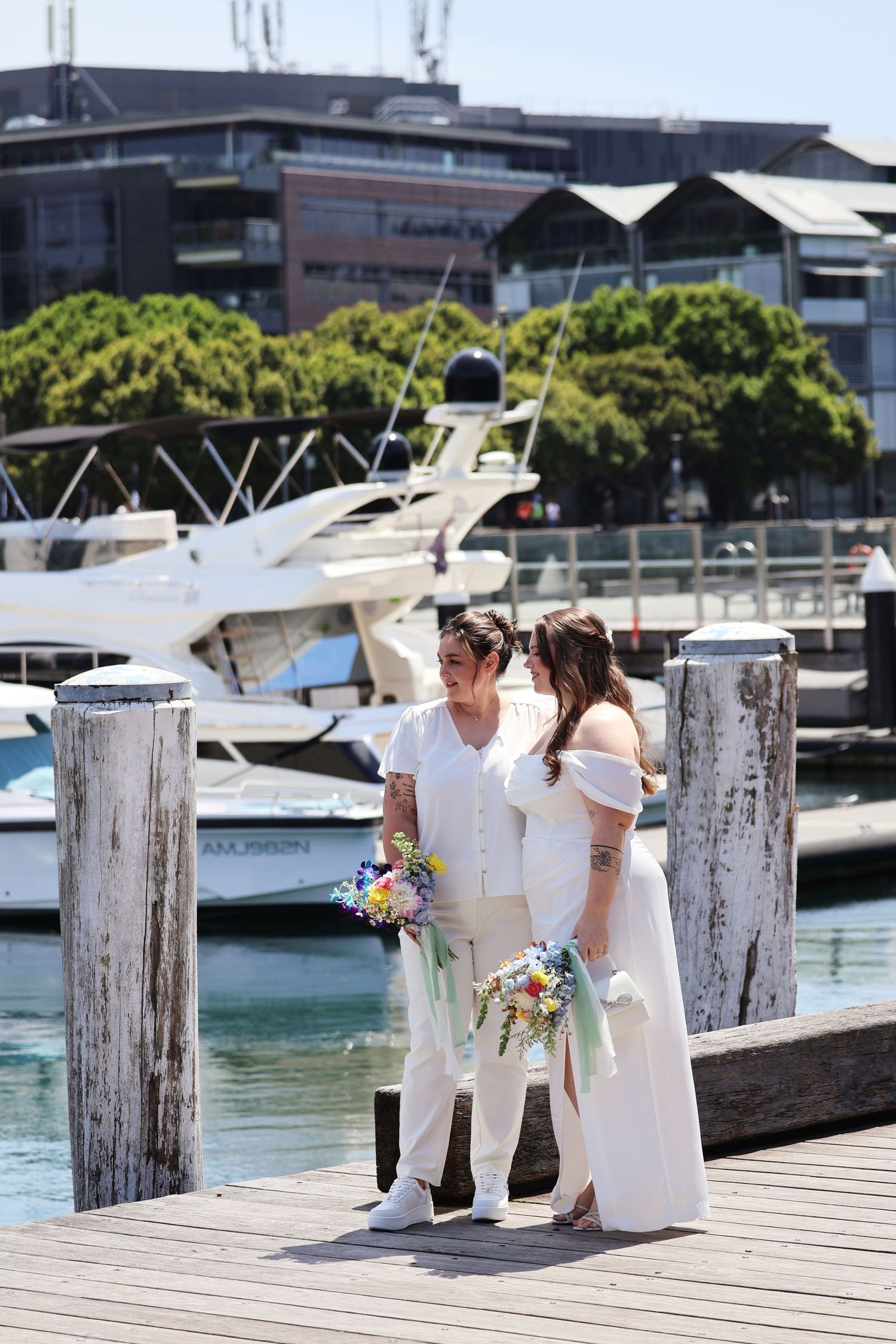 Same-sex couple in white wedding attire share an intimate moment on a marina dock with yachts behind.