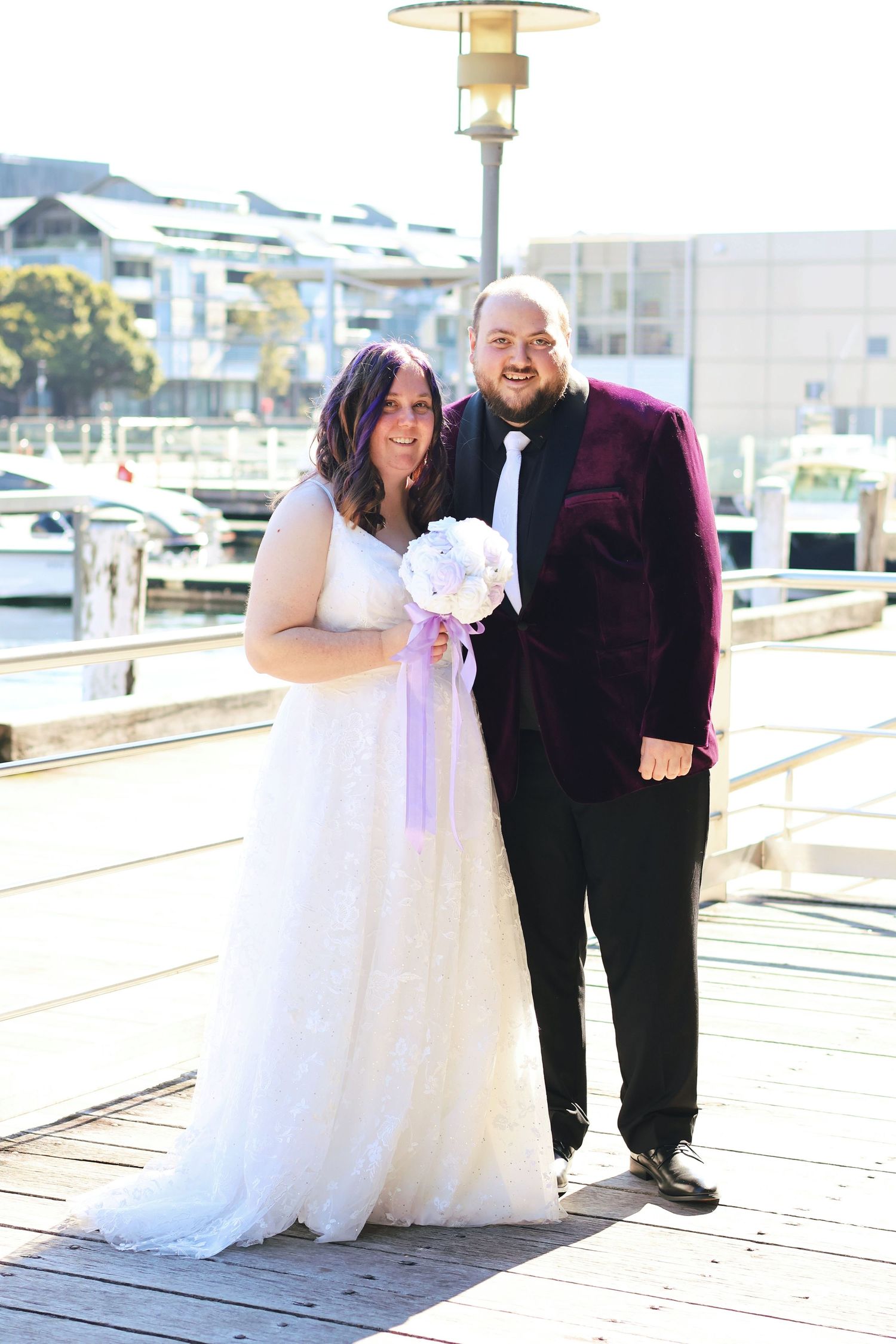 Bride in white lace gown and groom in burgundy velvet suit pose together on a sunny marina boardwalk.