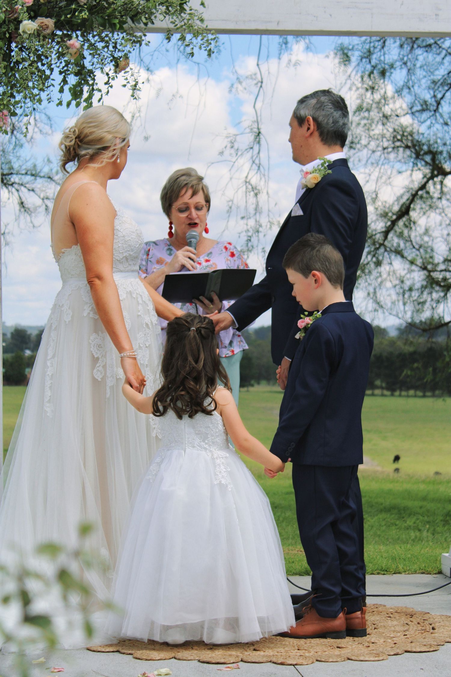 Wedding ceremony outdoors with bride, groom, two children, and officiant under floral arch on green lawn.