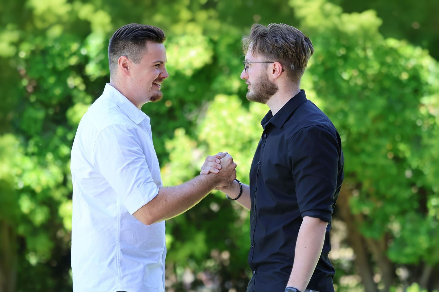 Two young men smiling and shaking hands outdoors surrounded by green trees on a sunny day.