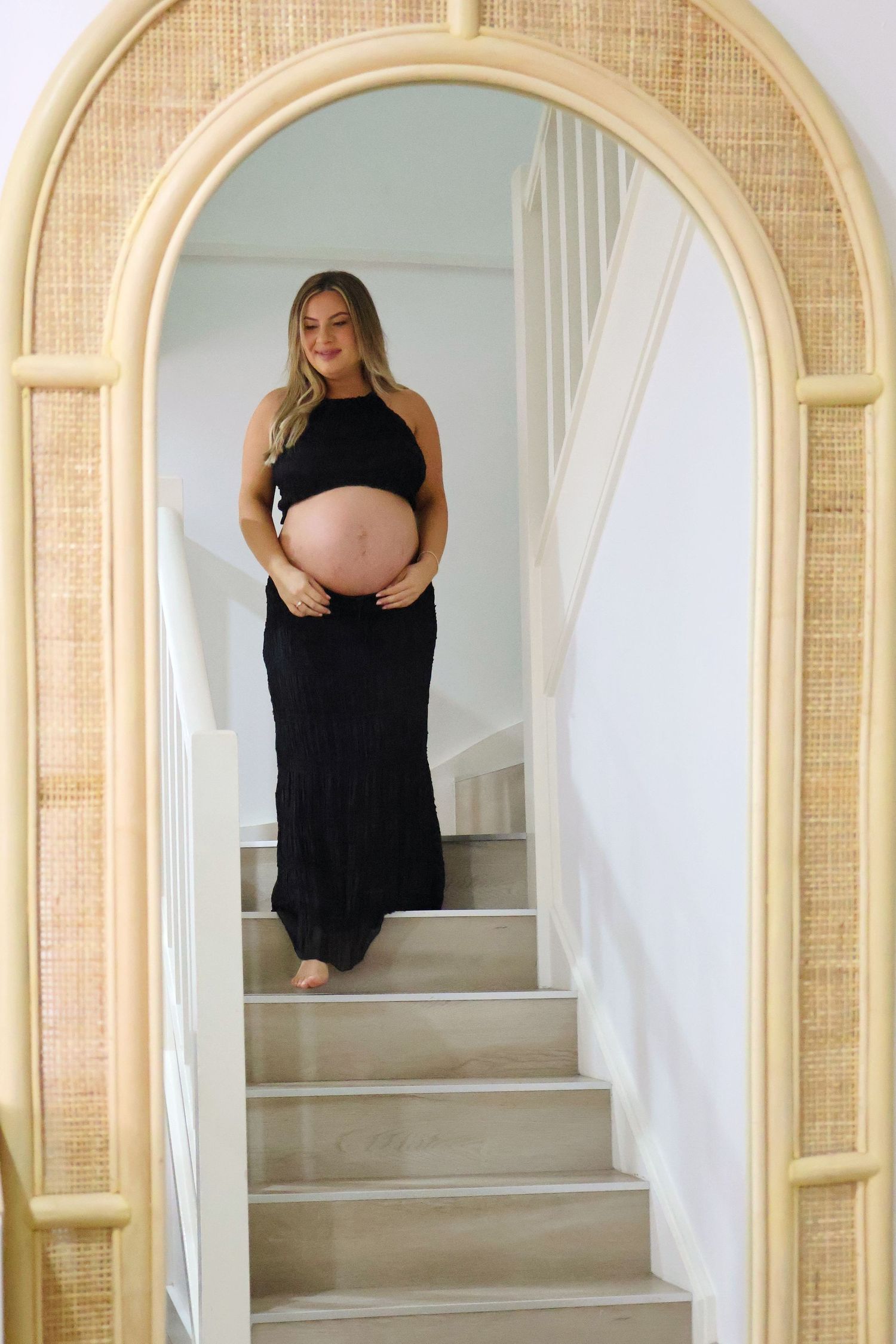 Pregnant woman in black maxi dress cradling baby bump while standing on stairs framed by arched rattan mirror.