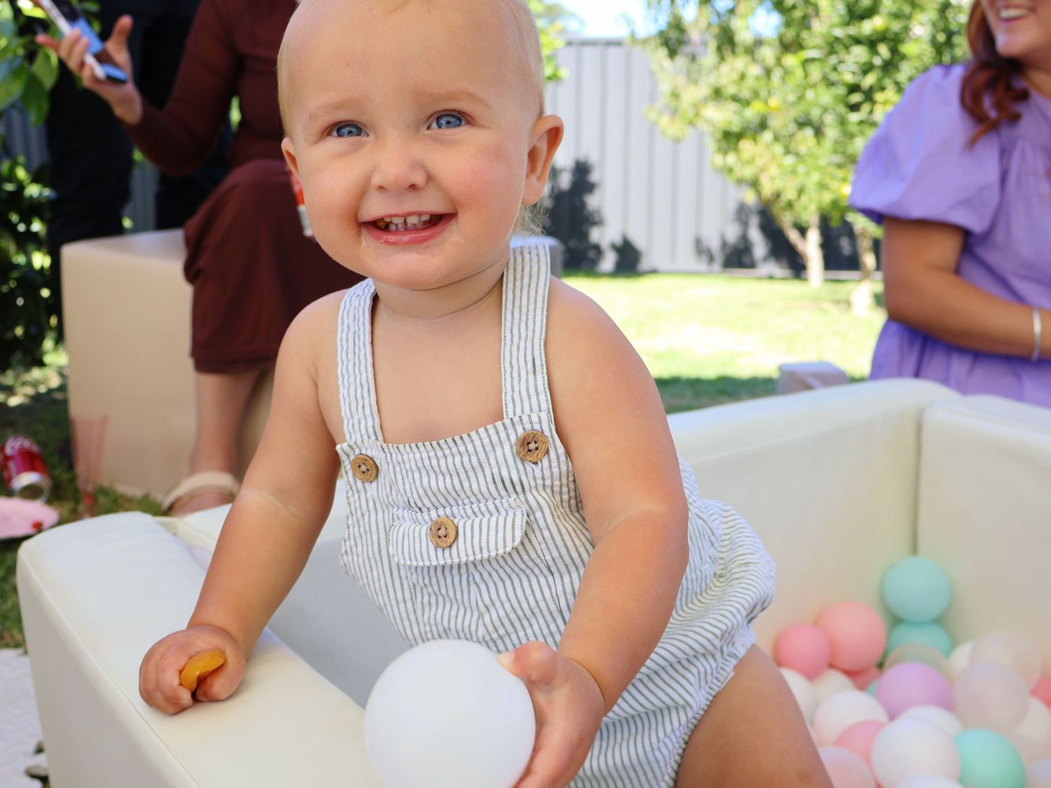 Happy baby boy sitting in a ball pit holding a white ball, smiling at an outdoor birthday party.