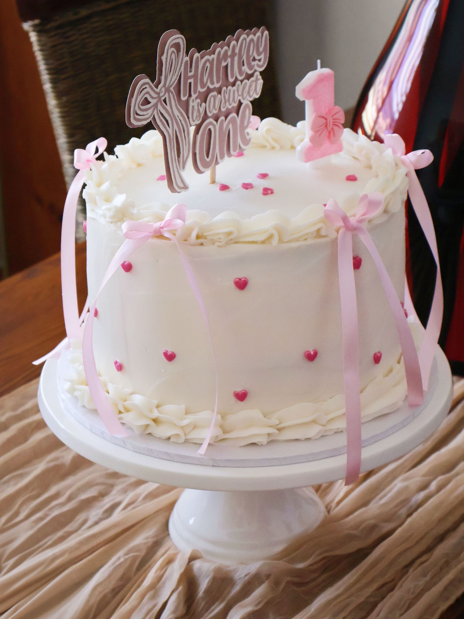 Elegant white first birthday cake with pink hearts, ribbons, and Happy Birthday One topper on a cake stand.