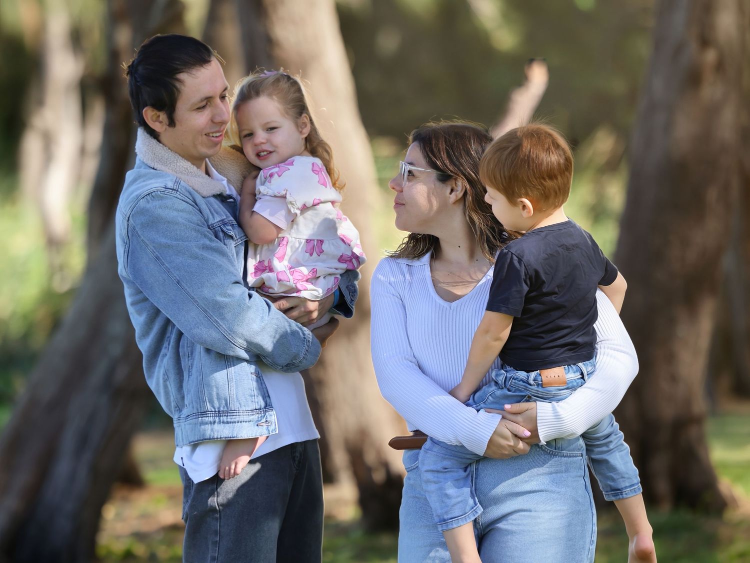 Happy young family with two toddlers smiling together among trees in outdoor park setting.