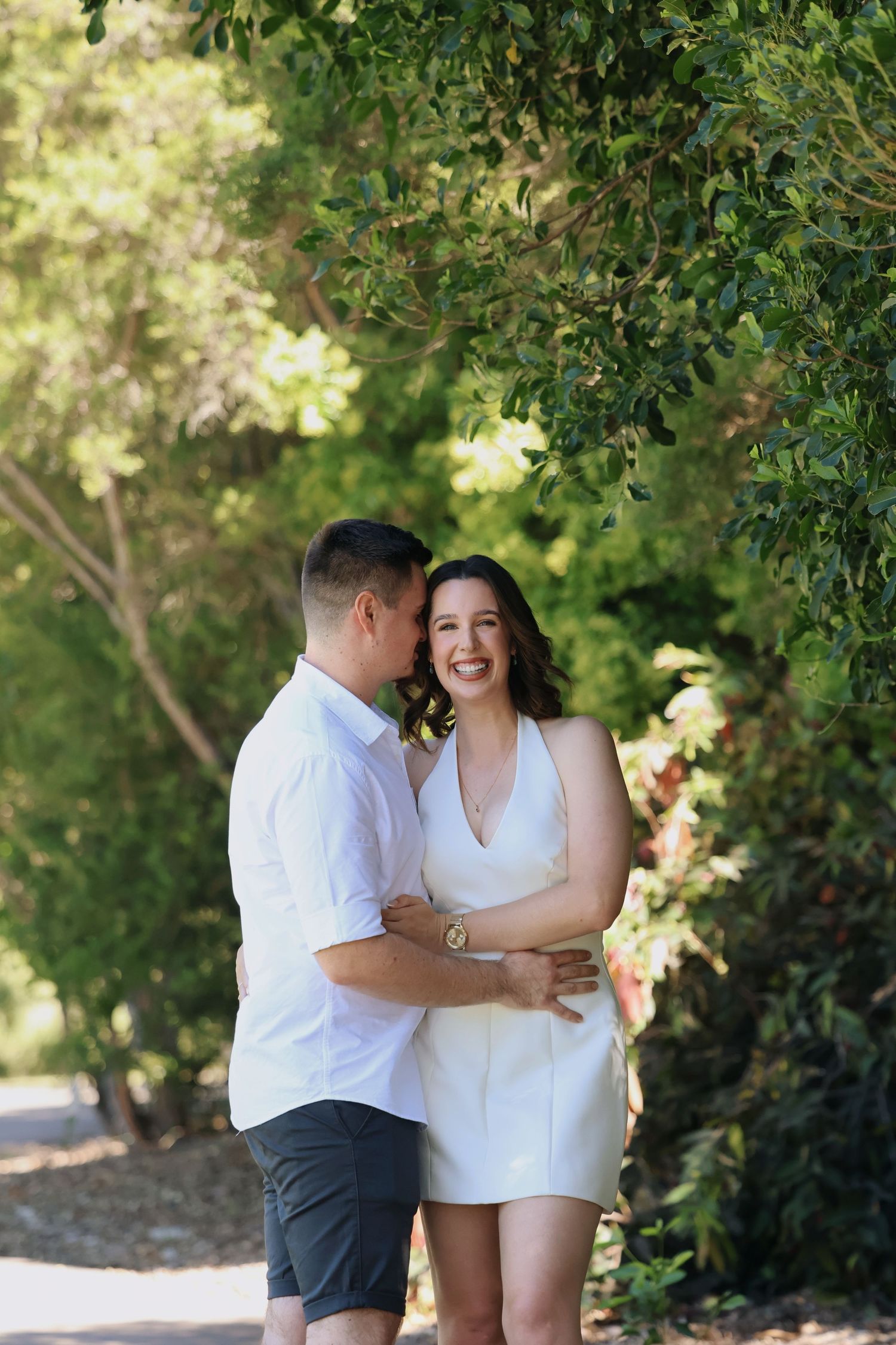 Smiling couple in white outfits embracing on a sunny garden path with green trees.