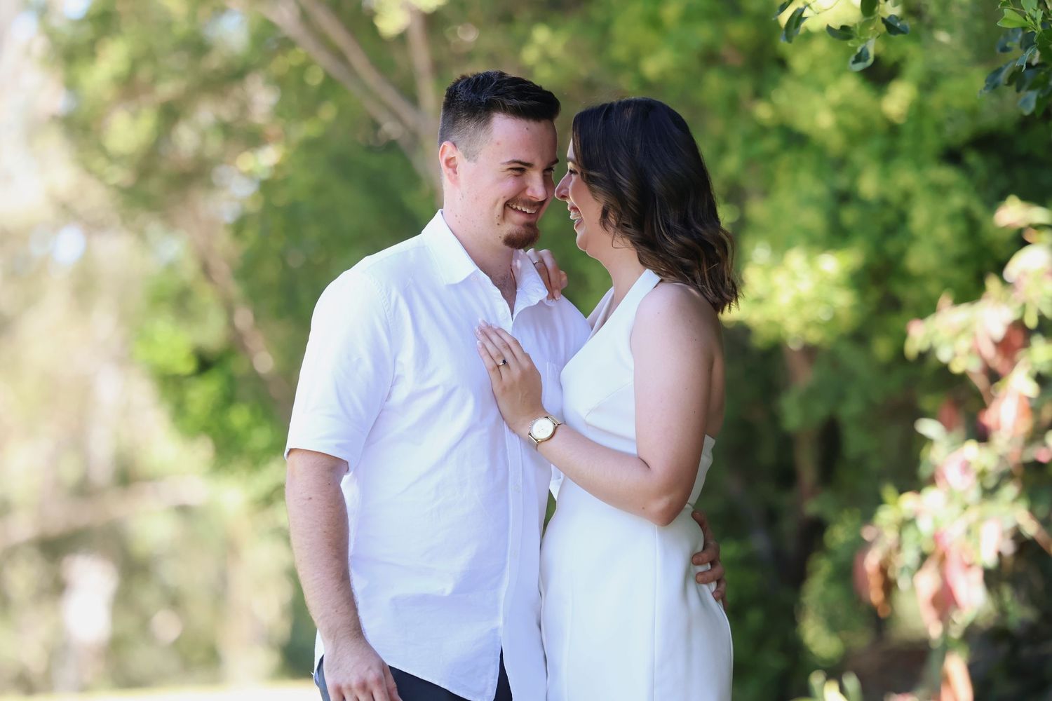 A couple shares a romantic moment outdoors, smiling at each other surrounded by lush green trees.