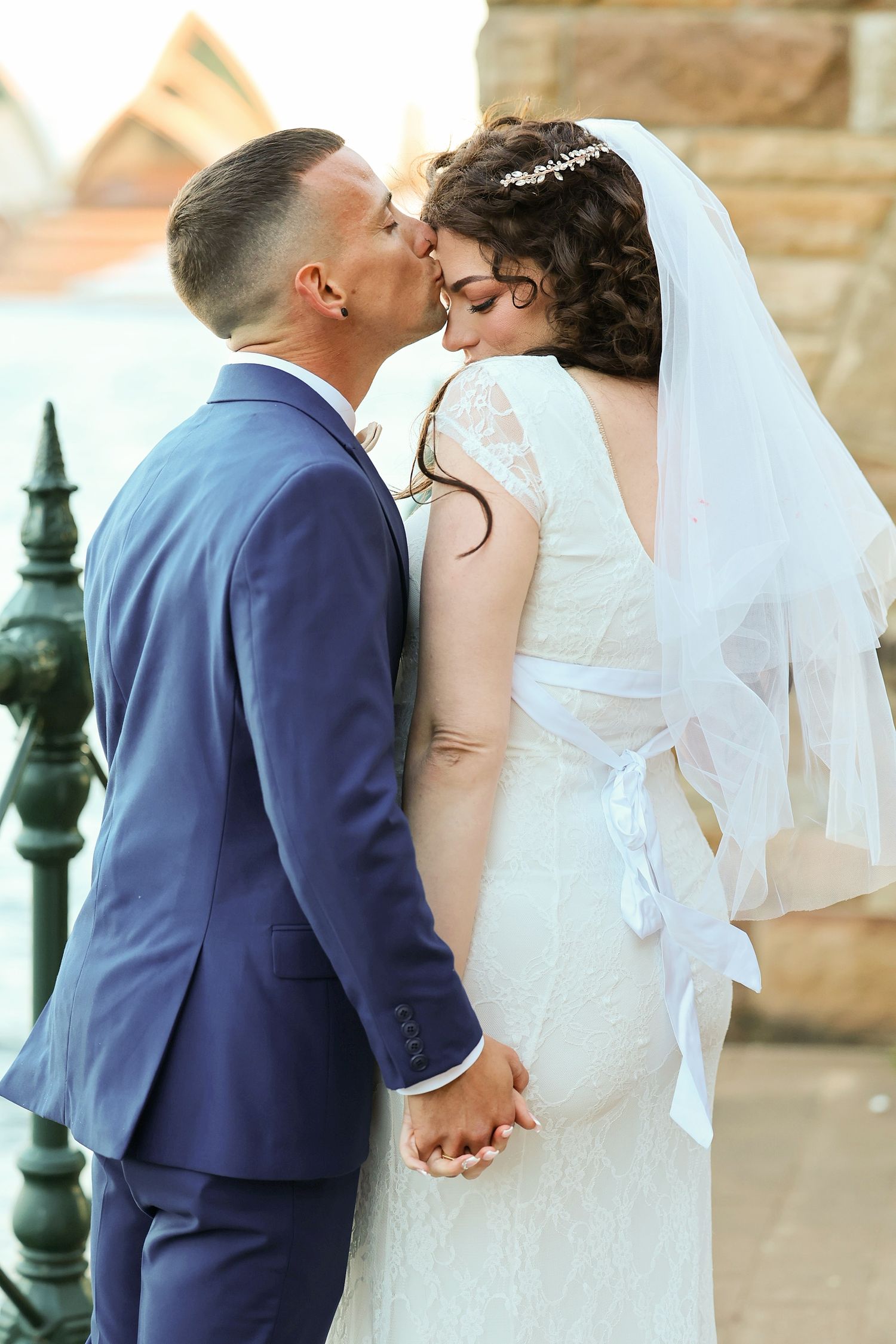 Bride and groom sharing a kiss near iron fence with Sydney Opera House in background.