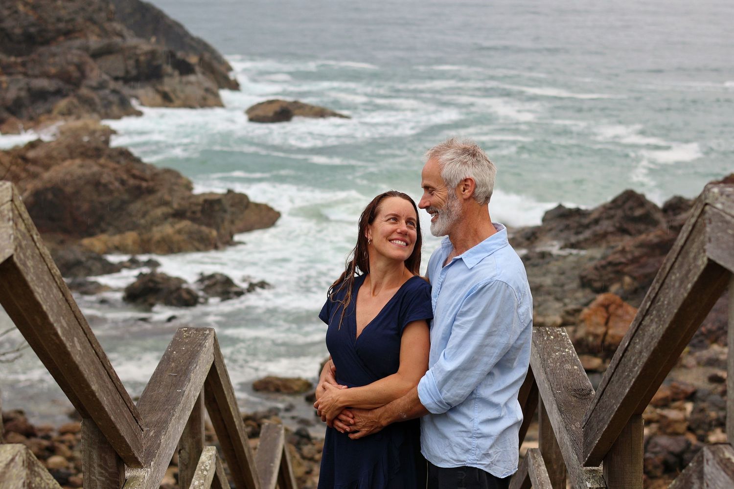 Couple embracing and smiling at each other on coastal stairs with rocky ocean waves in the background.