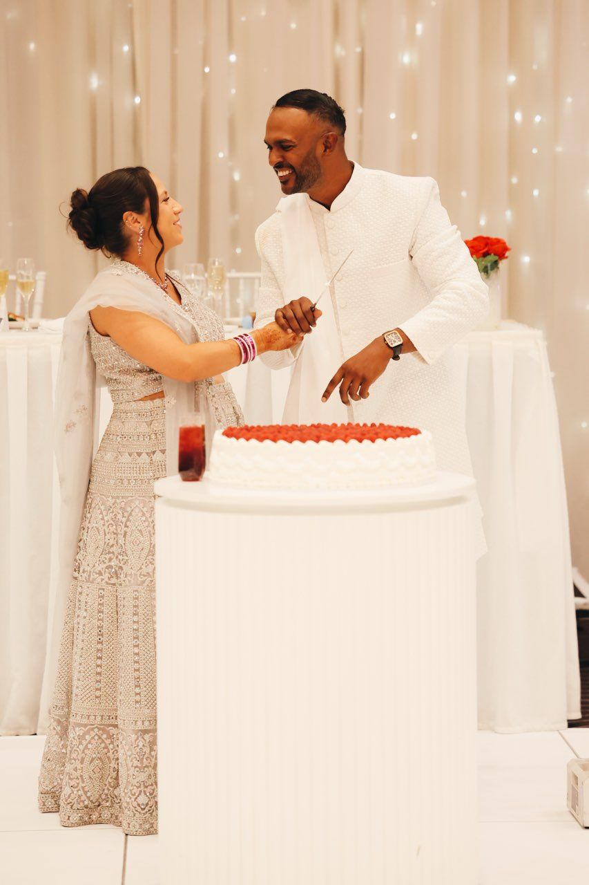 Couple cutting wedding cake together, bride in silver lehenga and groom in white suit, smiling at each other.