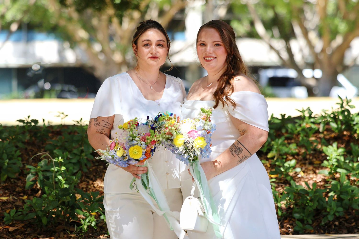 Two brides in white holding colorful bouquets smiling outdoors on sunny wedding day.