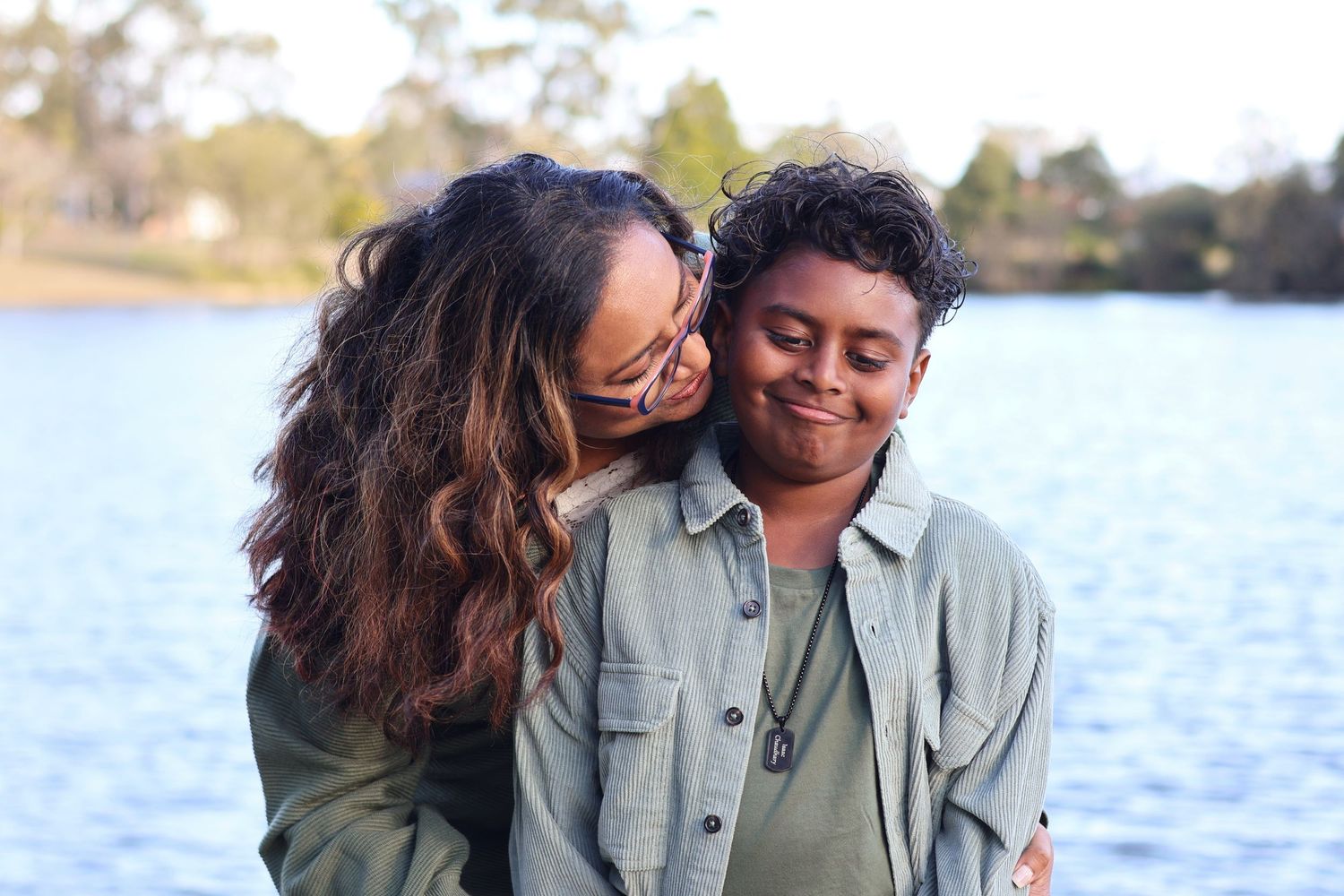 Two women share a tender moment embracing and smiling by a serene lake outdoors.