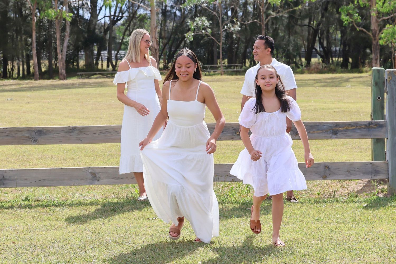 Happy family in white outfits running and playing together in a green field near a wooden fence.