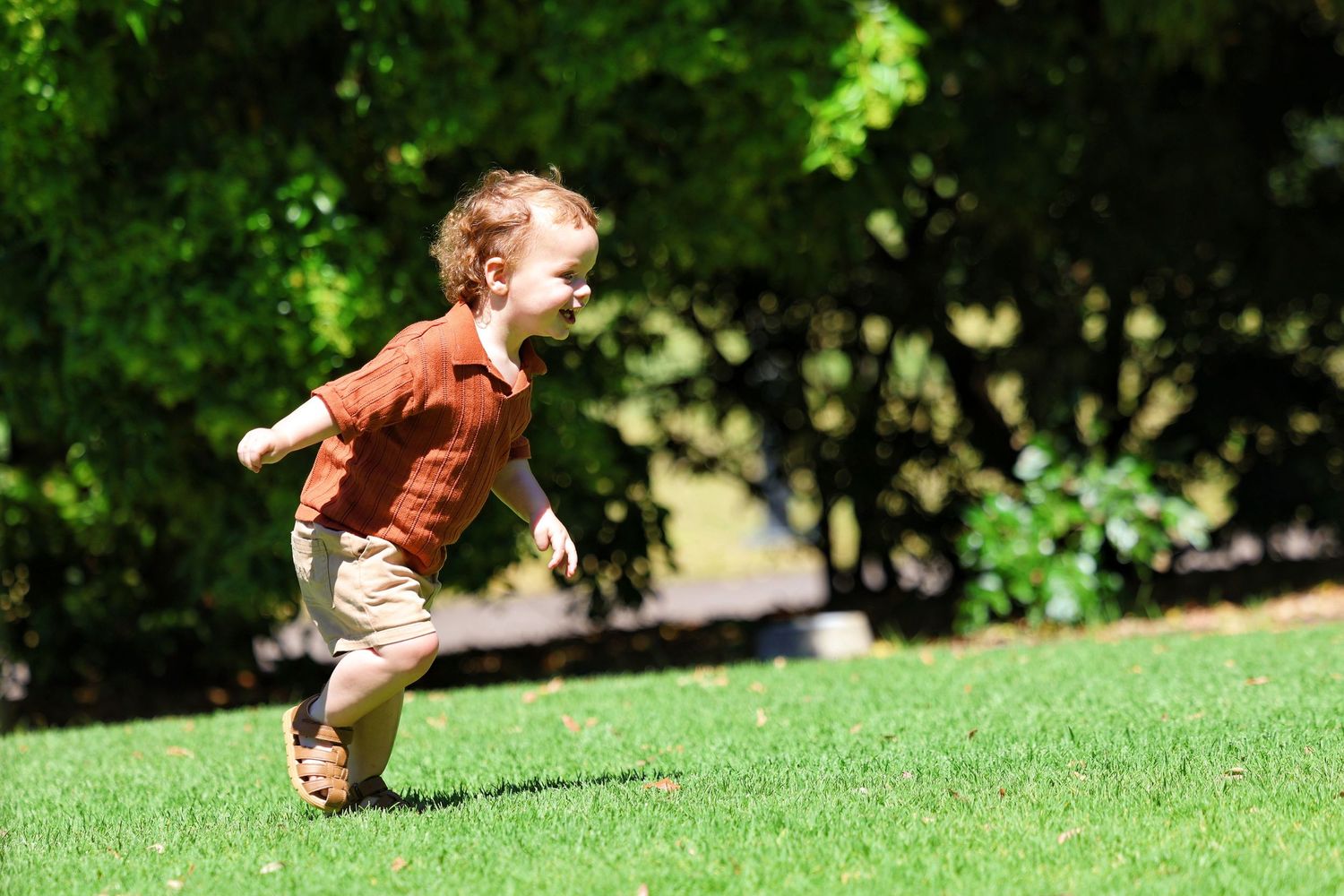 Energetic toddler in a rust shirt running joyfully across a lush green lawn.