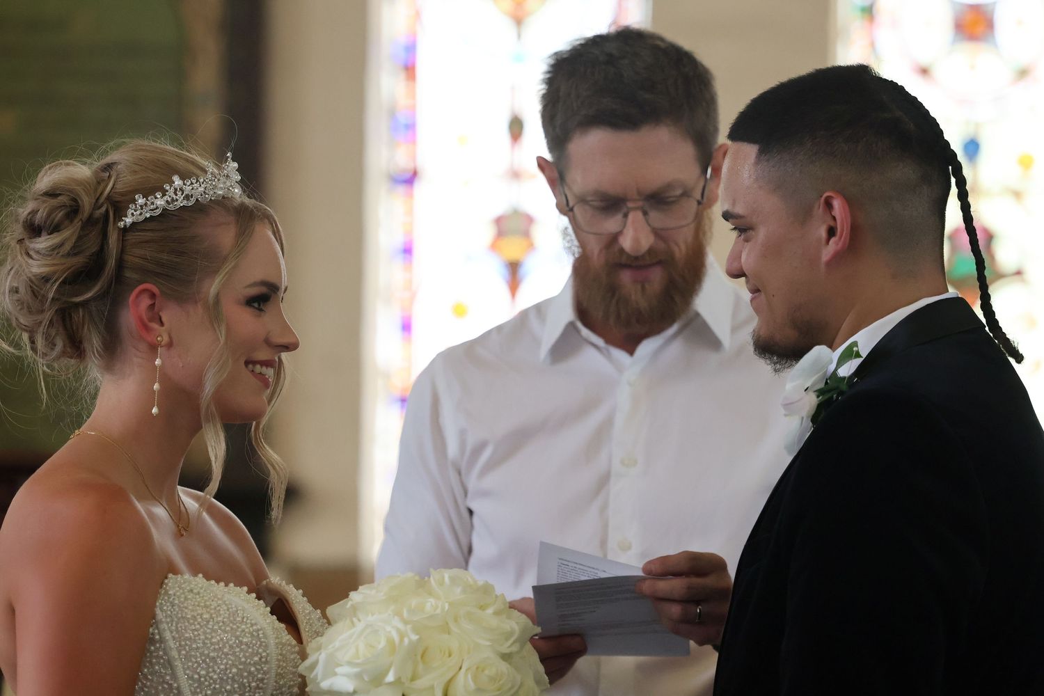 Bride and groom exchanging vows as bearded officiant reads from paper in church setting.