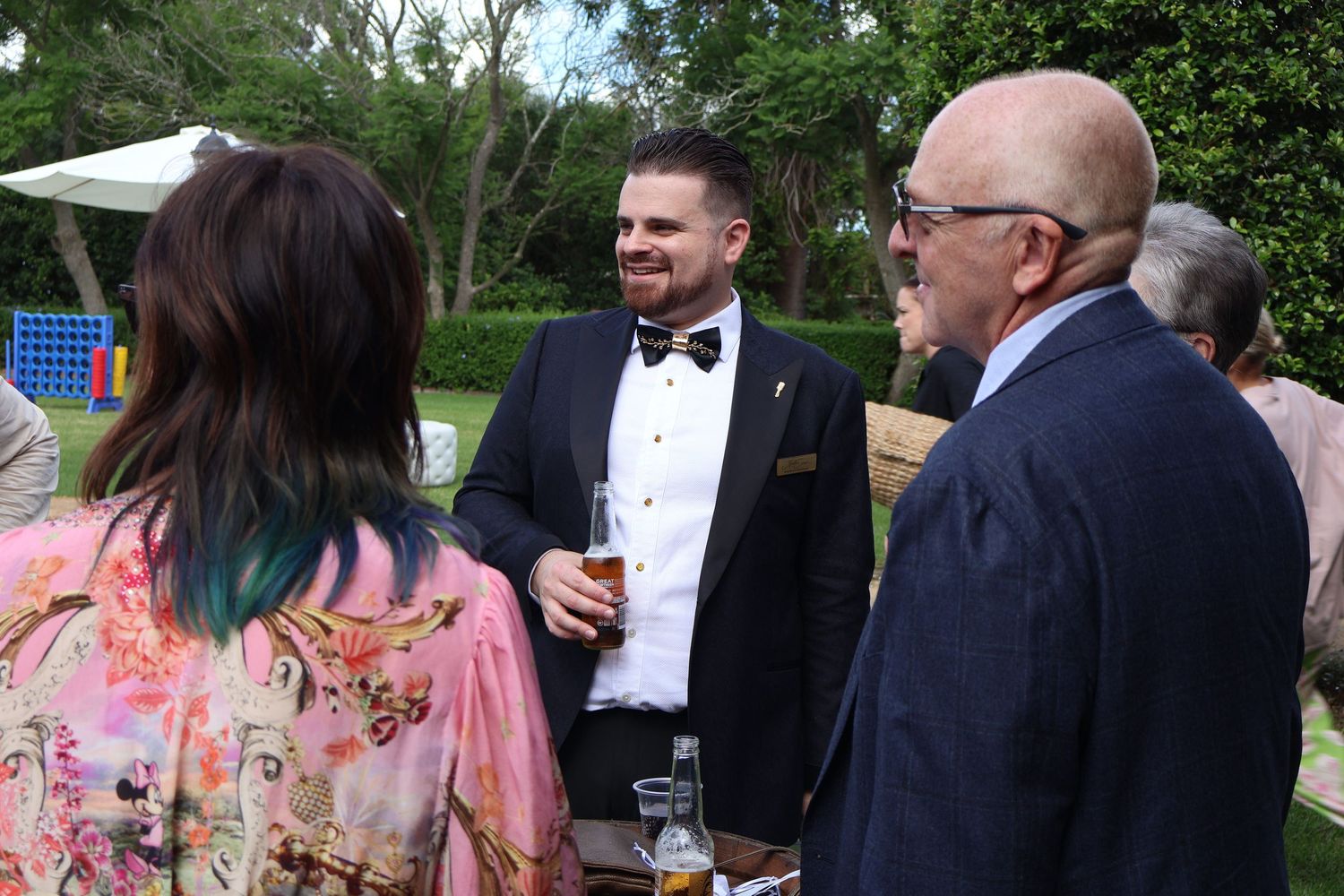 Man in black tuxedo with bow tie socializing at an outdoor garden wedding reception event.