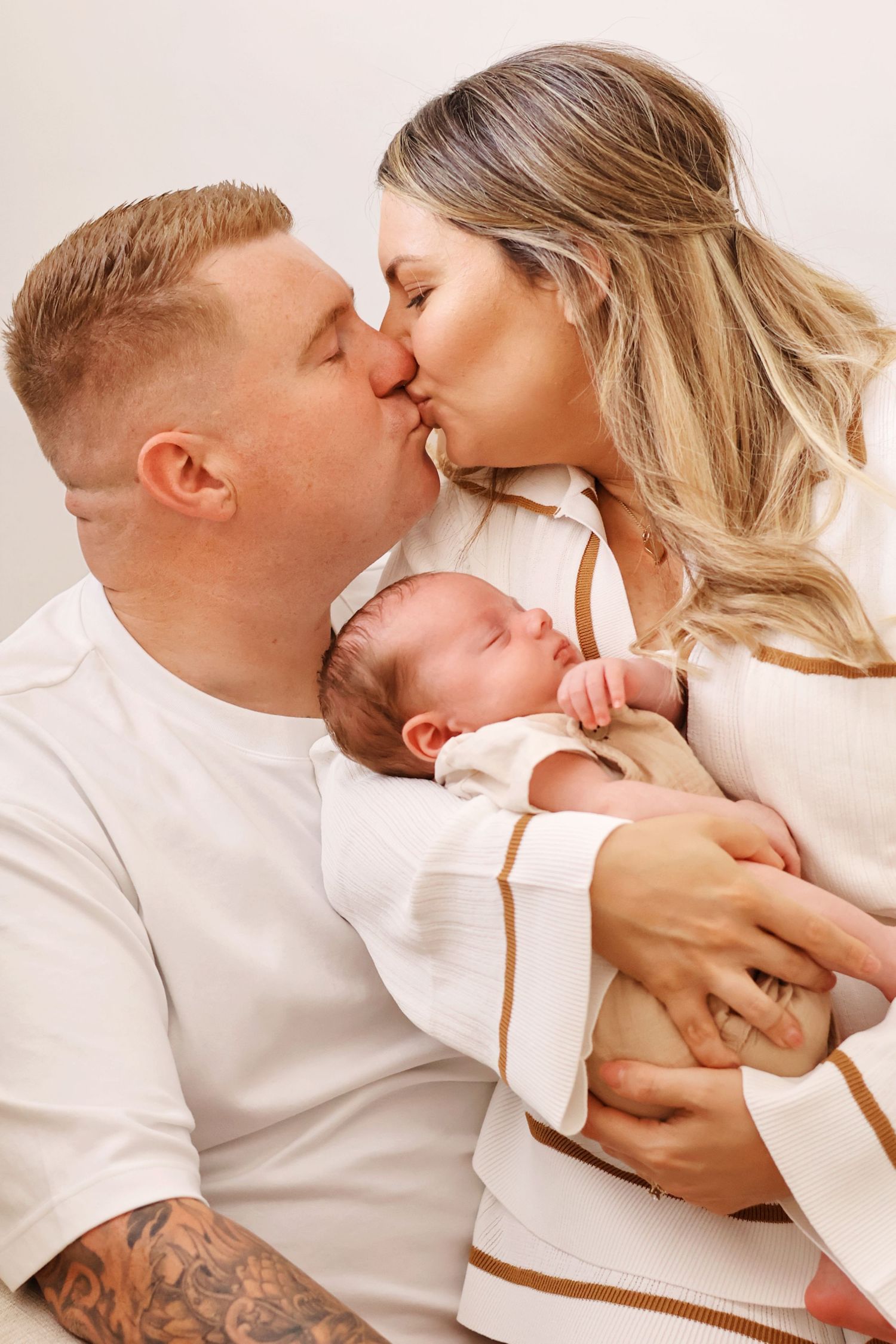 Happy couple kissing while holding their newborn baby dressed in white and tan outfit.