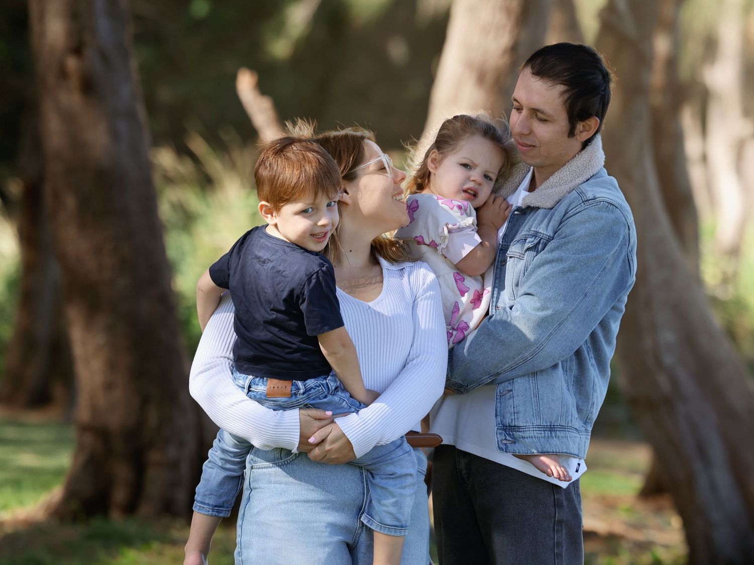 Family of four with two young children enjoying a moment together in a sunlit forest park.