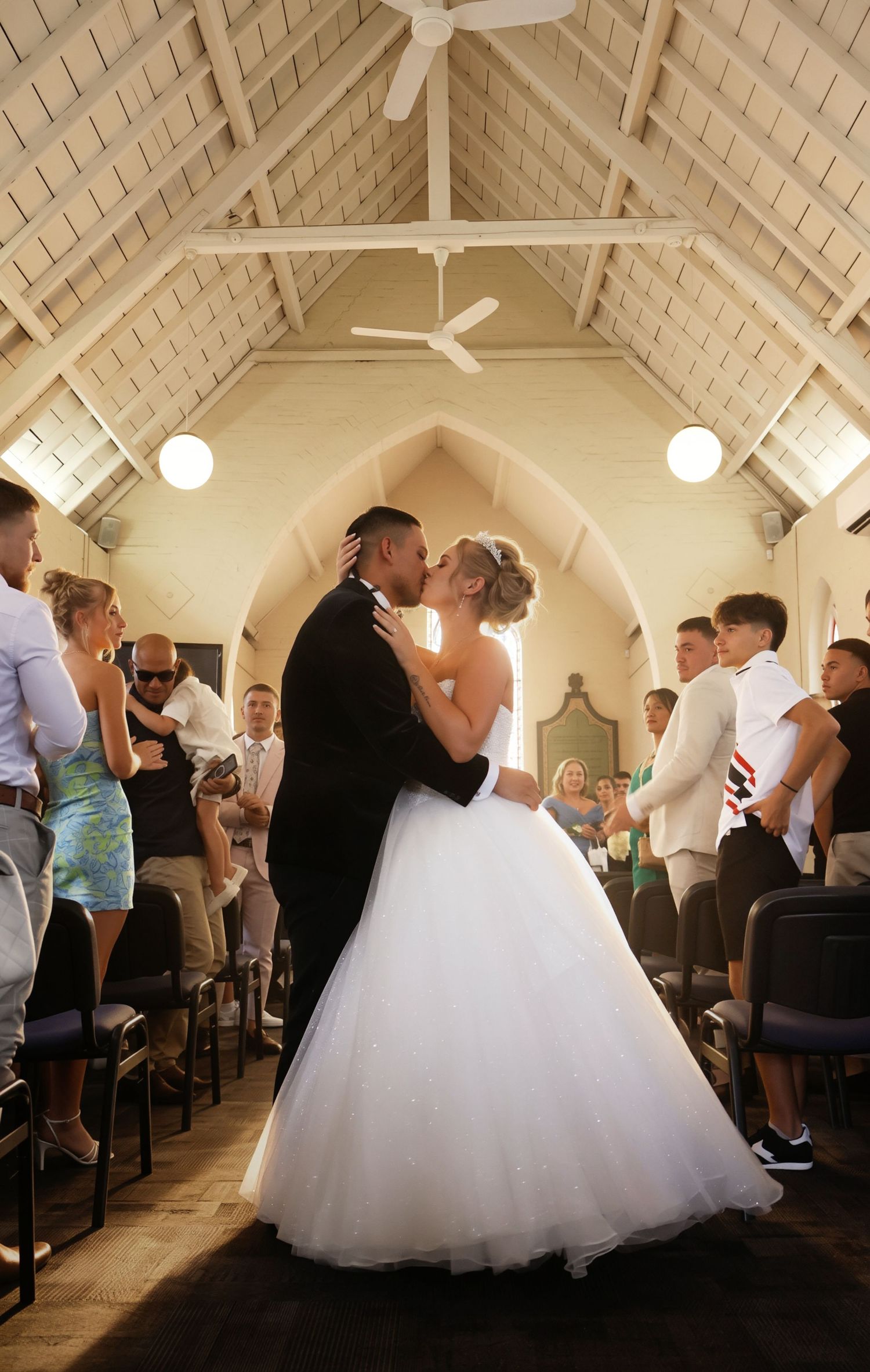 Bride and groom share their first kiss at the altar in a charming white timber chapel with guests applauding.