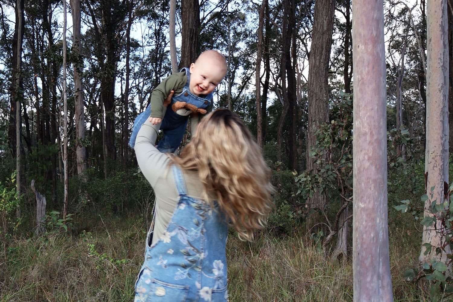 Woman lifting laughing baby overhead in a lush forest setting.