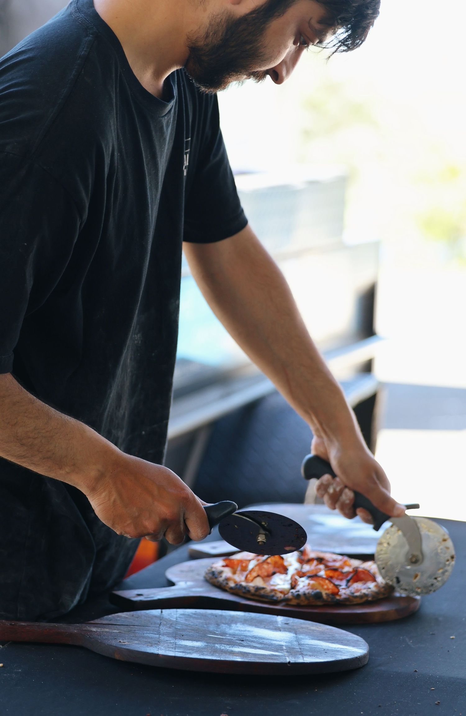 Bearded chef in black shirt slicing freshly baked pizza on a wooden board outdoors.