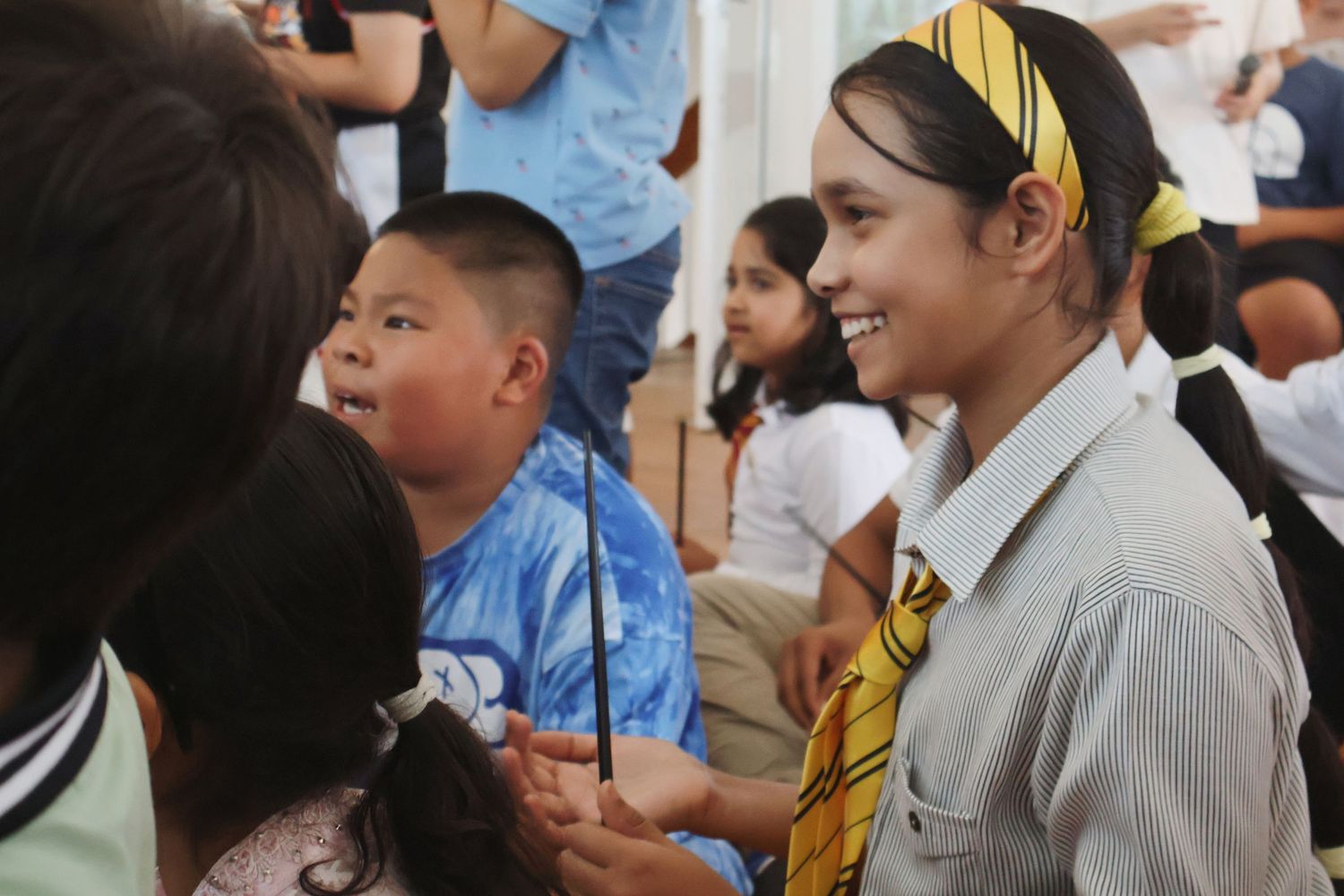 Smiling teenage girl in school uniform with yellow headband at a crowded indoor school event.