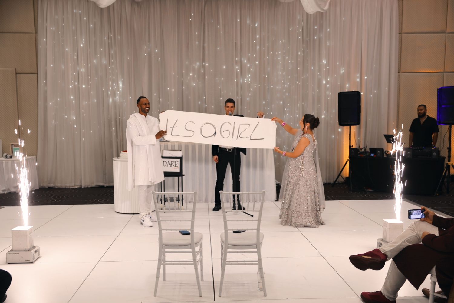 Three people hold a decorative sign on a white dance floor with sparkling backdrop at wedding reception.