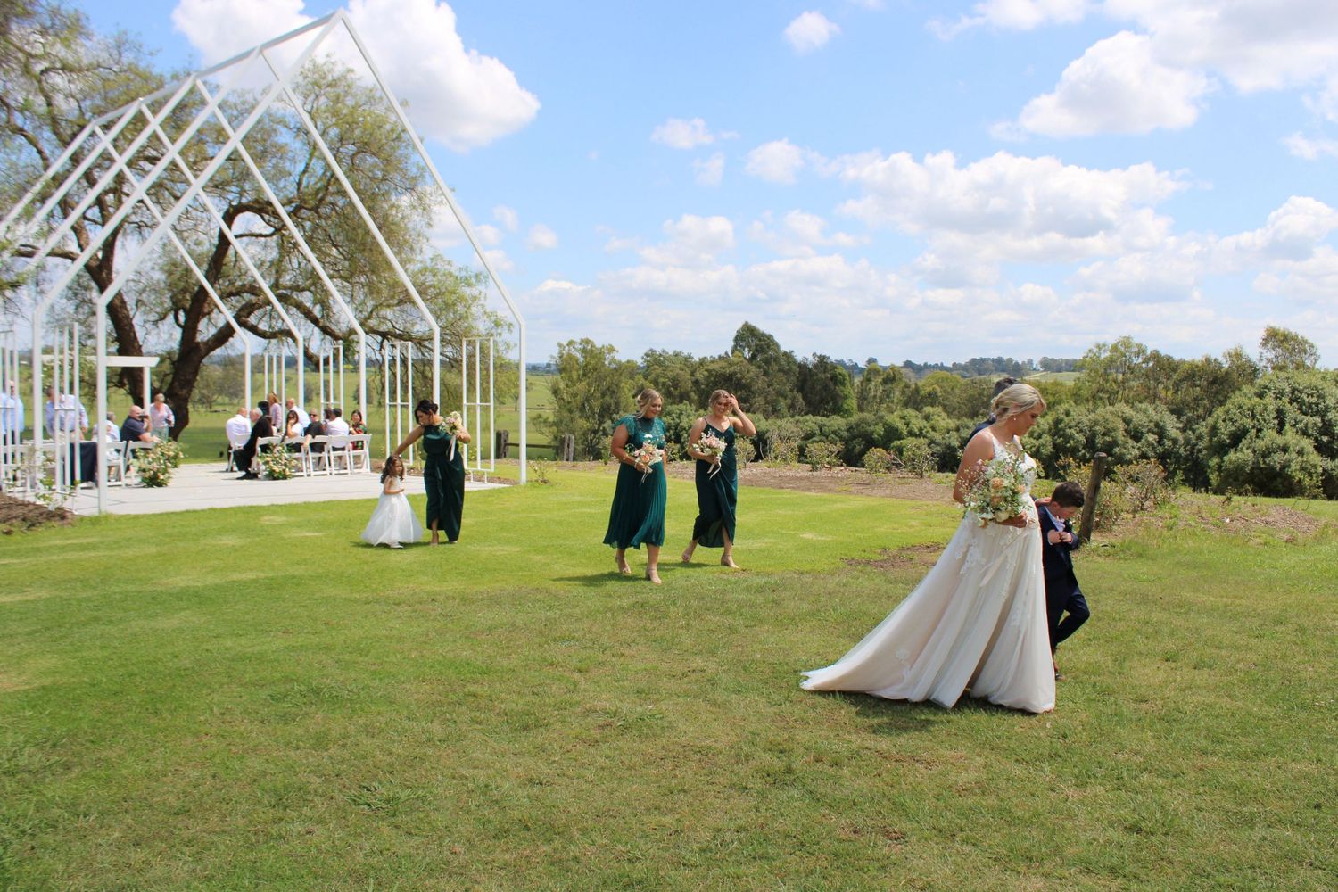Wedding party walking across lawn toward elegant glass greenhouse ceremony venue on sunny day.
