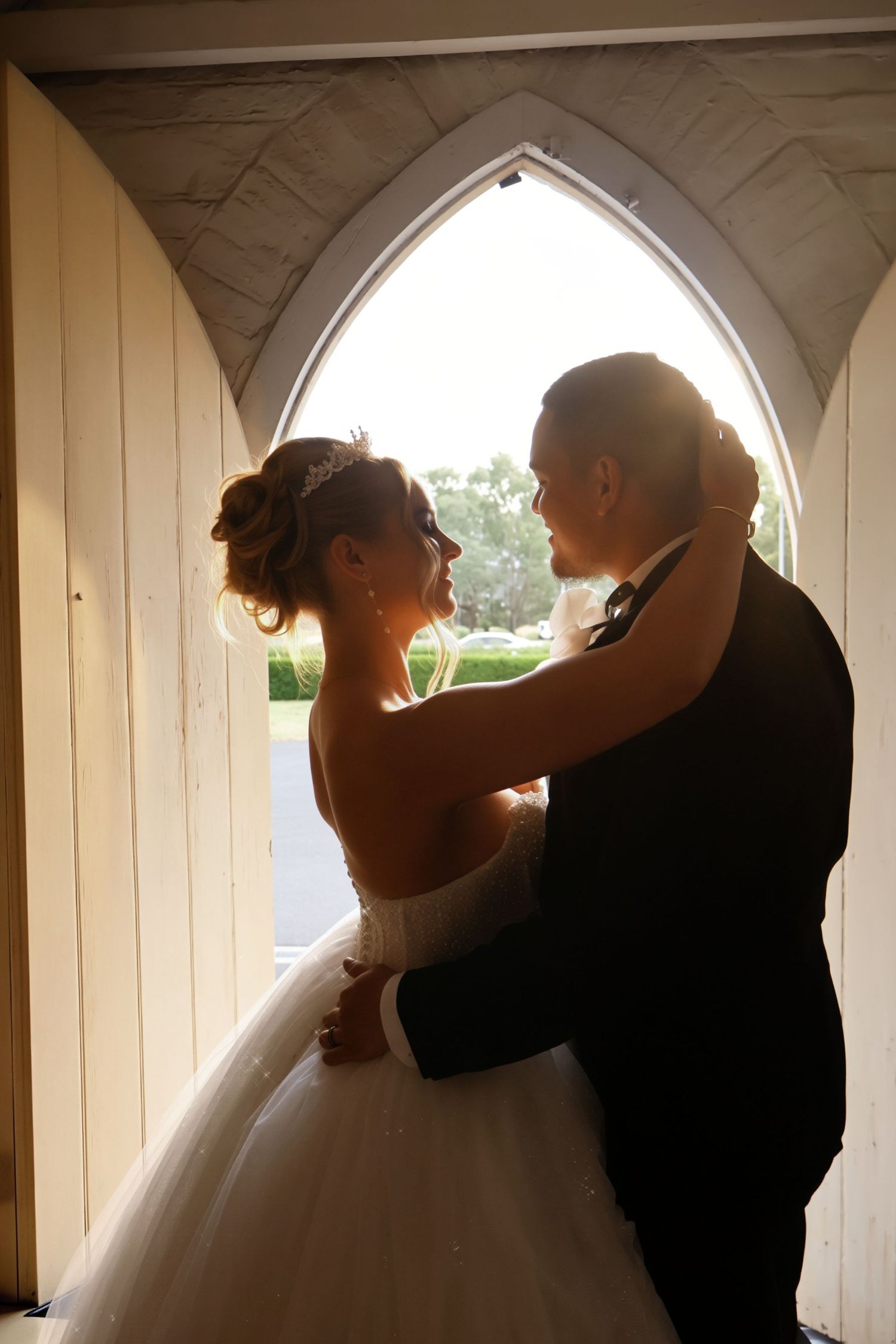 Bride and groom share a romantic embrace silhouetted against a Gothic arch window in a white chapel.