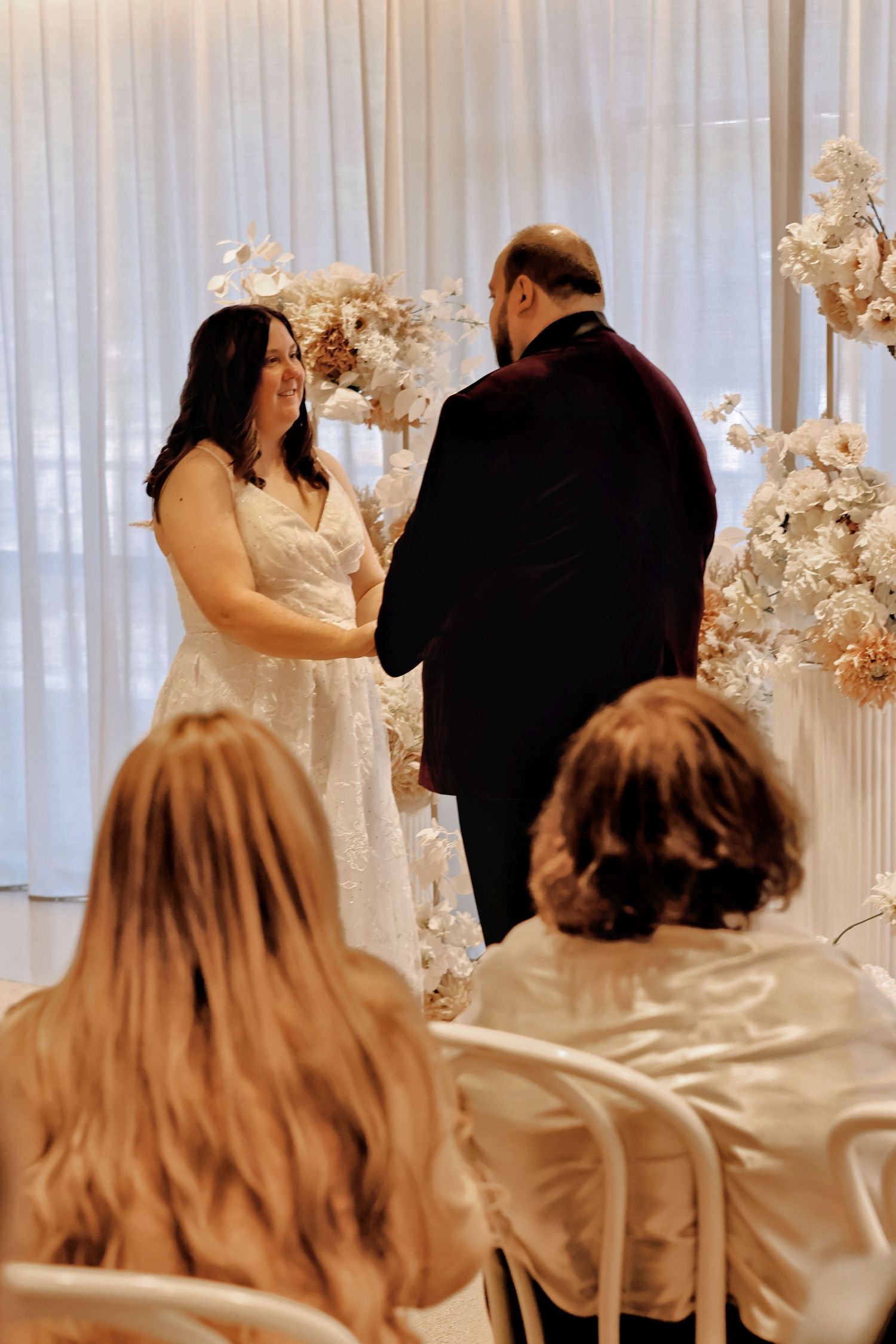 Smiling bride exchanging vows with groom at a white floral wedding ceremony with seated guests in foreground.