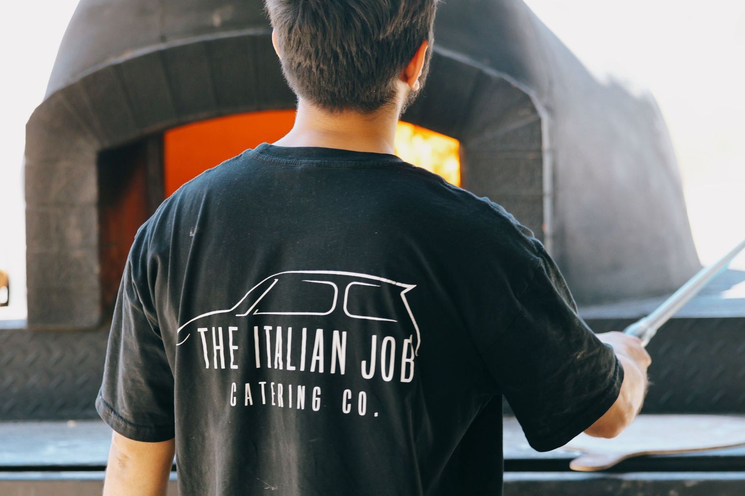 Man wearing The Italian Job Catering Co. black t-shirt standing in front of a wood-fired pizza oven.