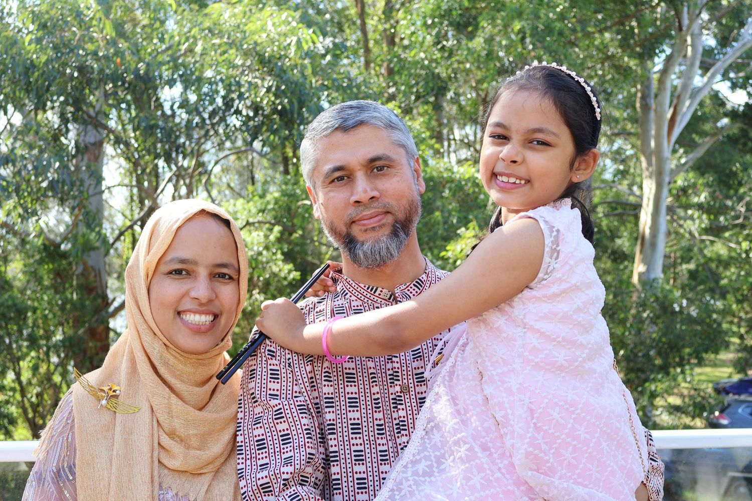 Muslim family portrait with mother in hijab, father with beard, and young daughter smiling outdoors in a park.