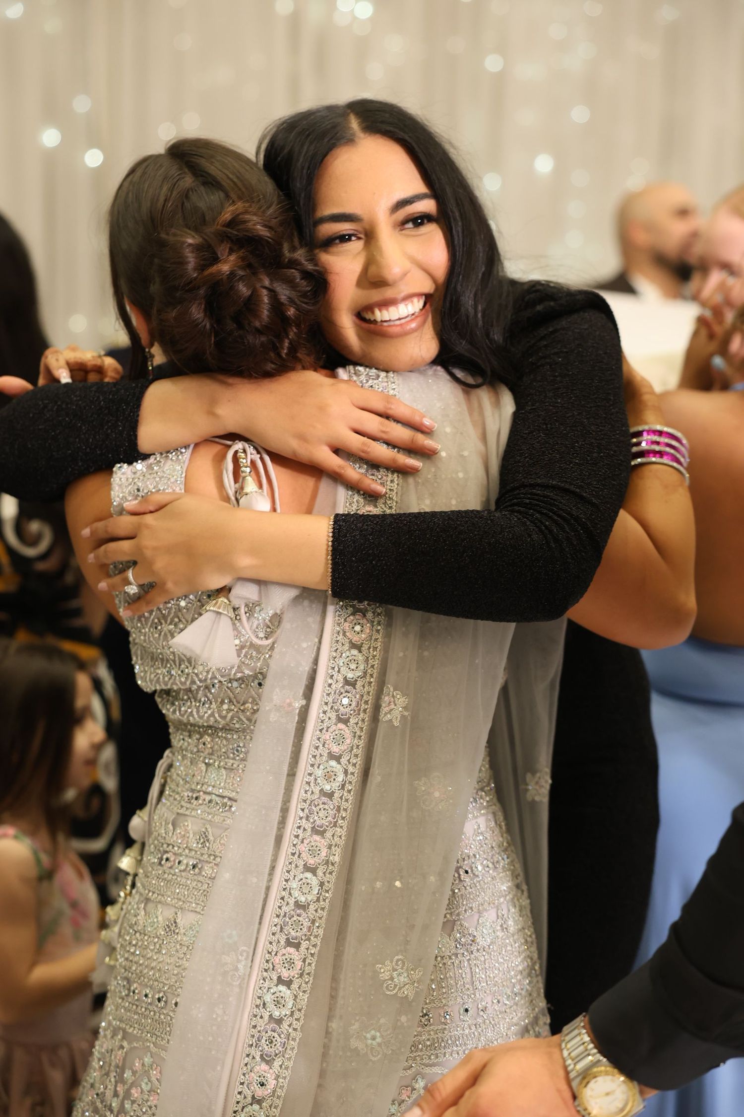 Woman in black top smiling while embracing guest in silver lehenga at wedding reception.