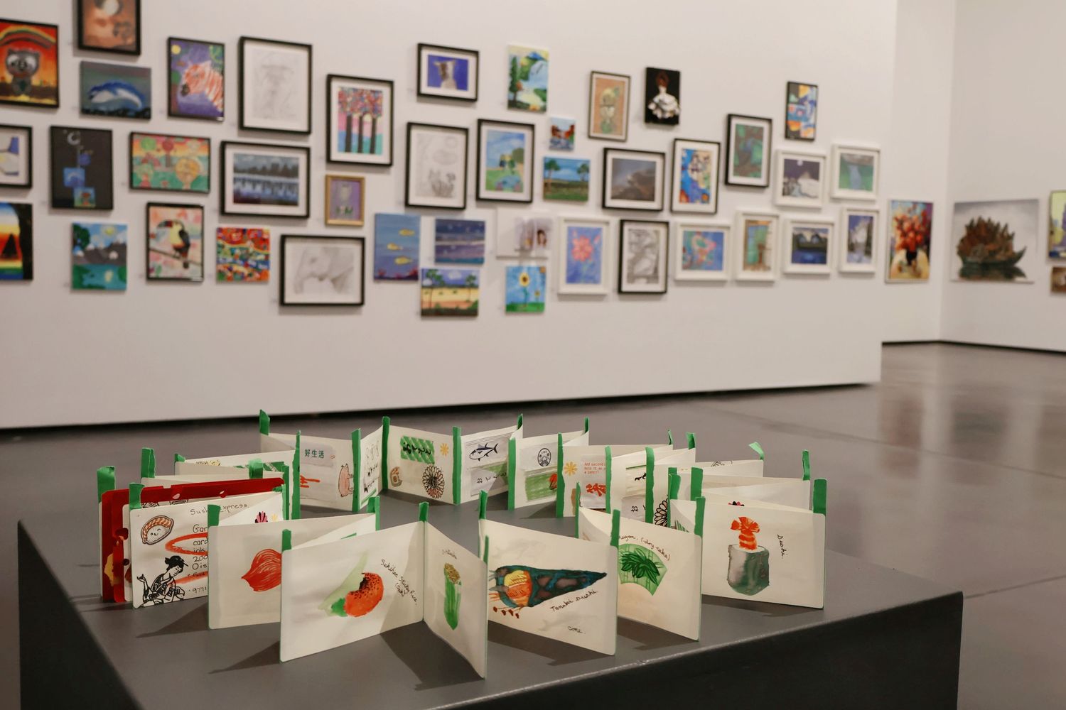 Colorful art exhibition wall filled with small paintings and drawings, with accordion books in foreground.