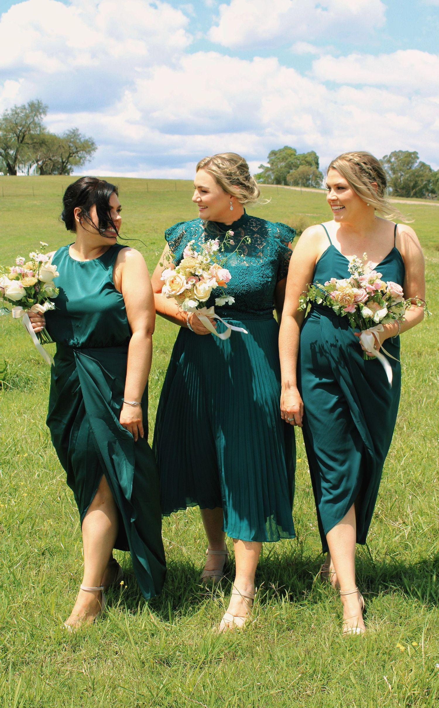 Three bridesmaids in emerald green dresses walking through green field holding pink and white bouquets.